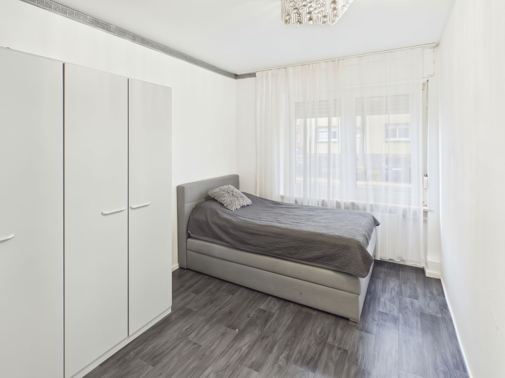 Helles Schlafzimmer mit grauem Bett, Kleiderschrank und Fenster mit weißen Vorhängen.