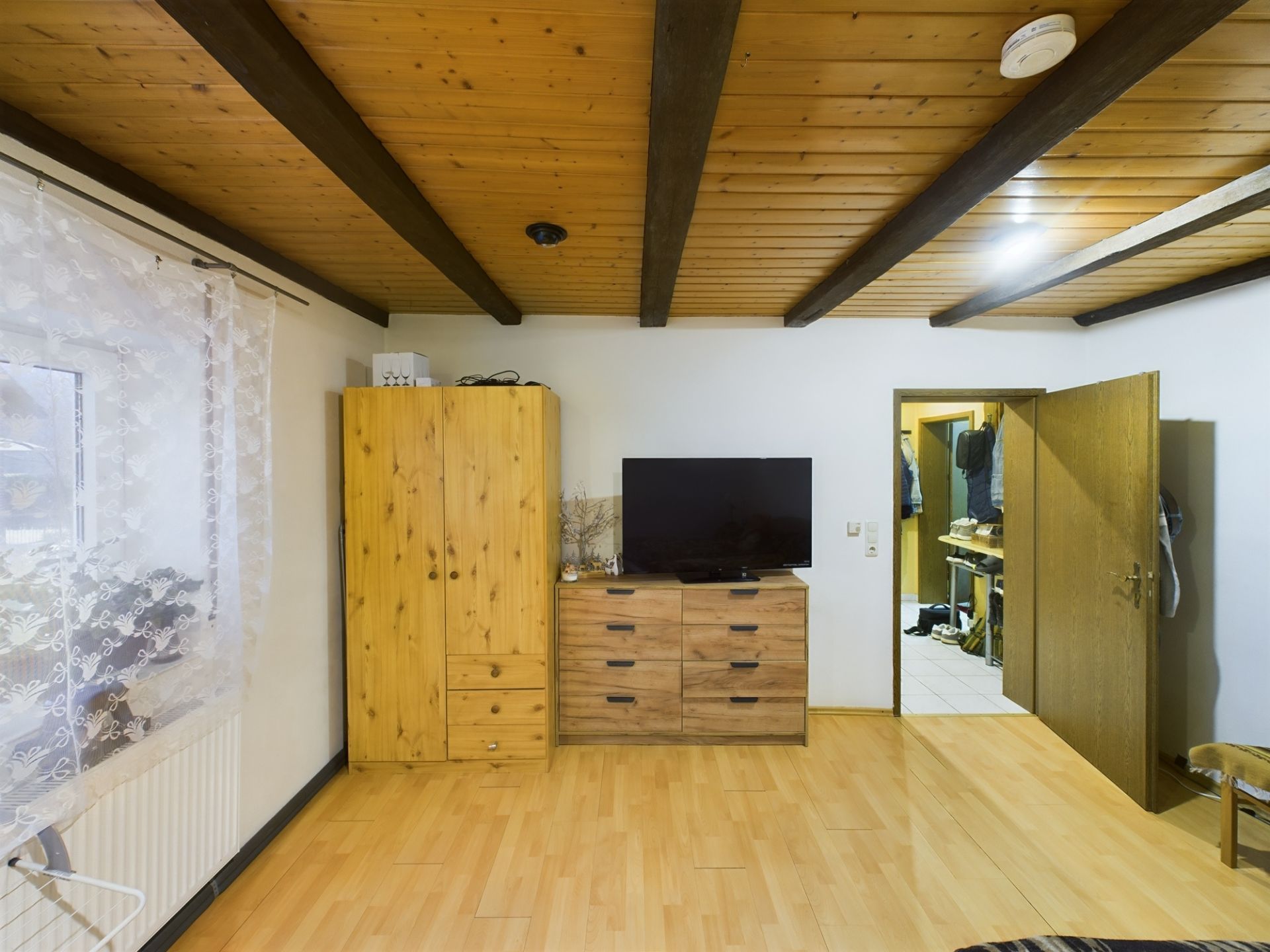 Wohnzimmer mit Holzdecke, Schrank, Kommode und Fernseher, Fenster mit Vorhang links.