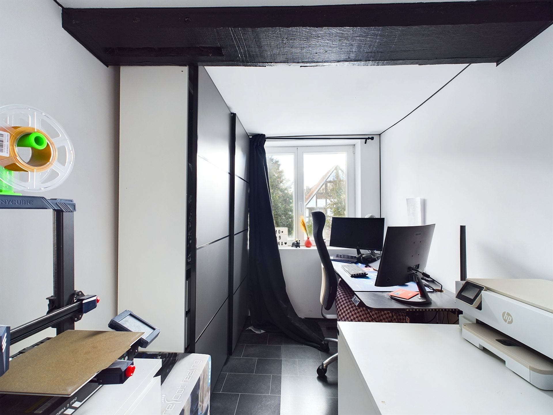 Schmaler Arbeitsraum mit Schreibtisch, Computer, Stuhl, Drucker und 3D-Drucker, Fenster im Hintergrund.