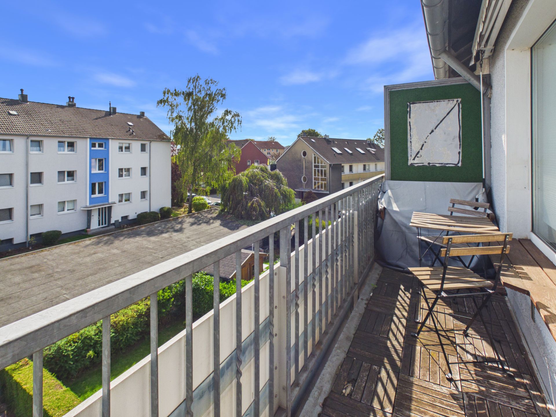 Balkon mit Holzboden, kleinem Tisch und zwei Stühlen, Blick auf Wohngebäude und Bäume.
