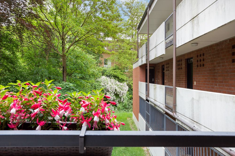 Blick von einem Balkon mit pinken Blumen auf einen begrünten Innenhof und Wohngebäude.