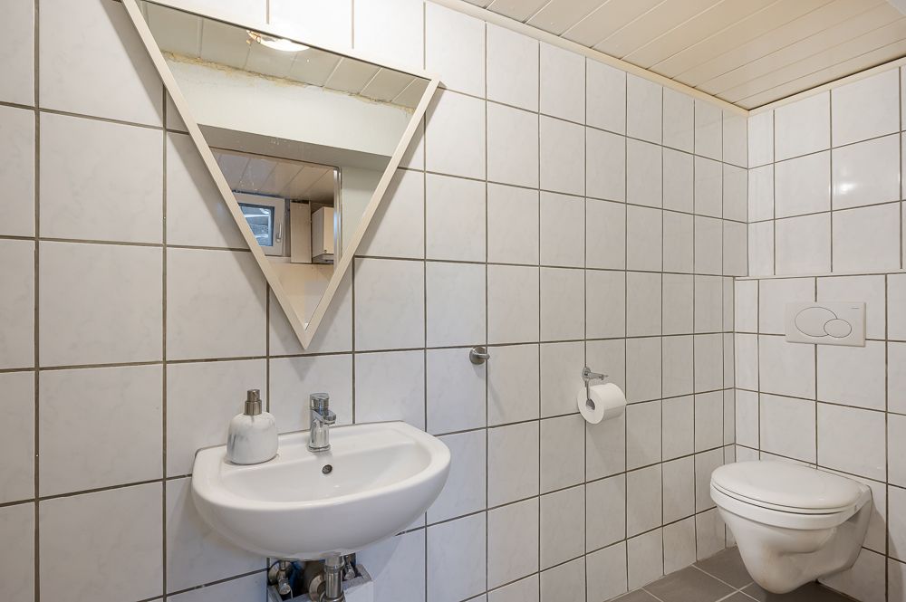 Kleines Bad mit Wand-WC, Waschbecken, Seifenspender und dreieckigem Spiegel.