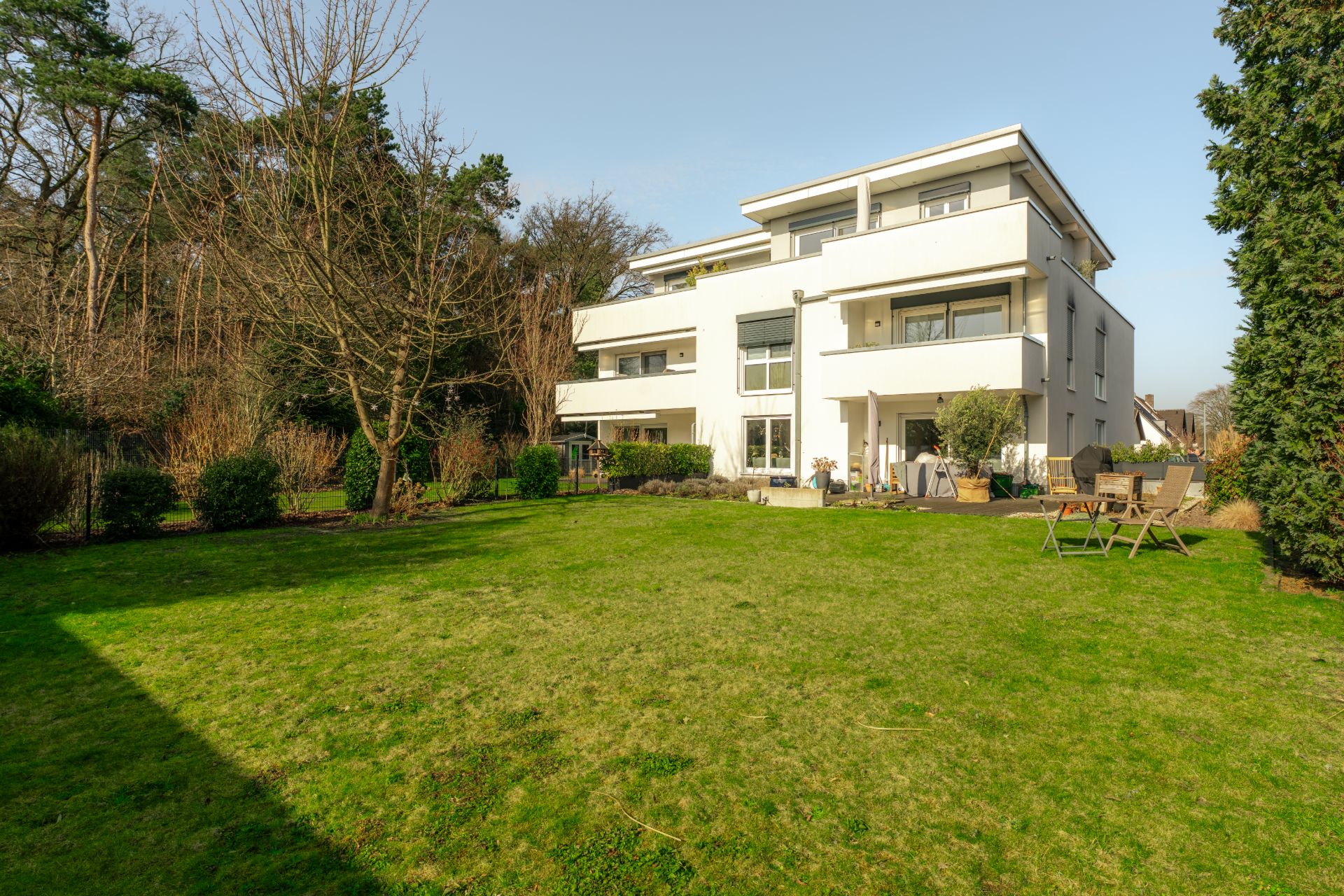 Modernes weißes Wohnhaus mit mehreren Balkonen, großer Garten mit Gartenmöbeln, Bäumen und Rasenfläche.