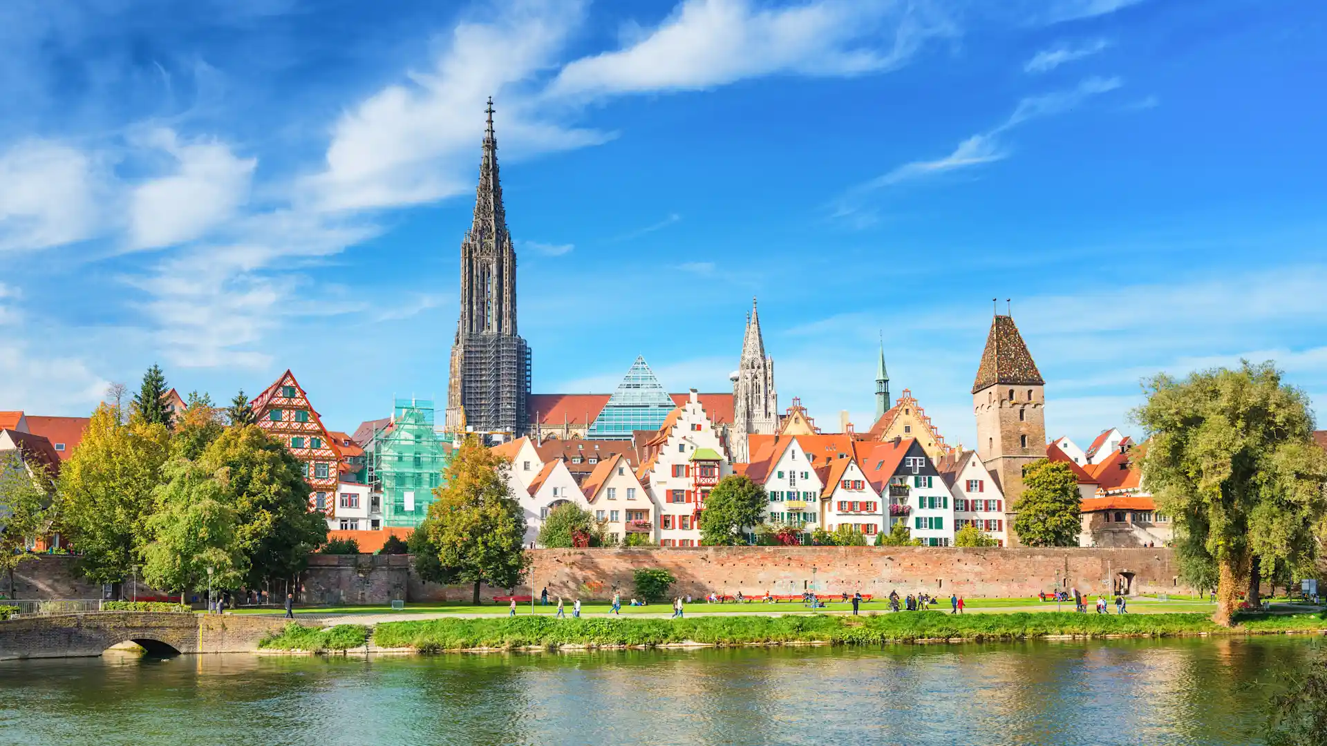 Bunte Häuser mit Giebeldächern am Fluss, dahinter Kirchtürme und Bäume.