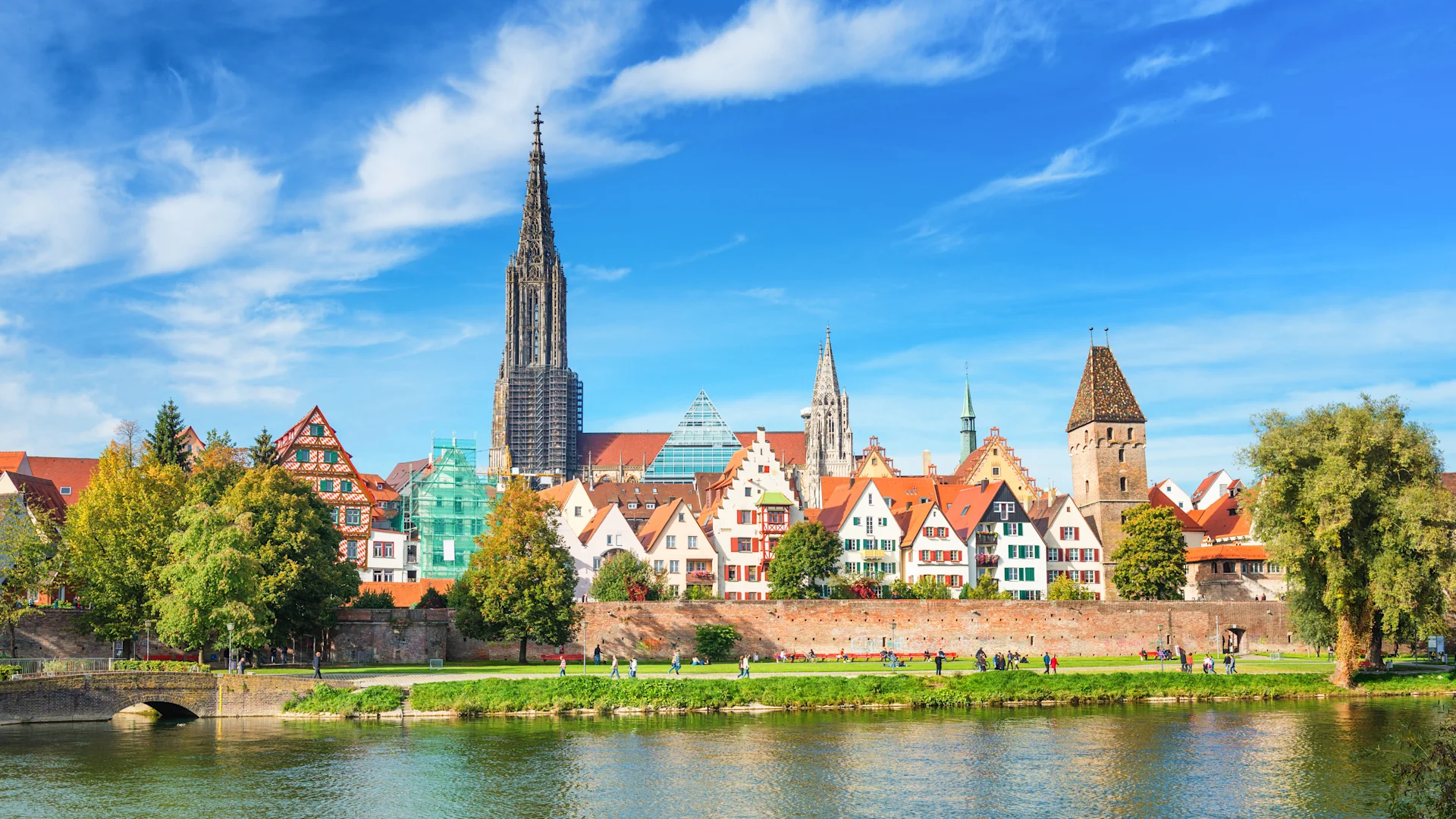Bunte Häuser mit Giebeldächern am Fluss, dahinter Kirchtürme und Bäume.