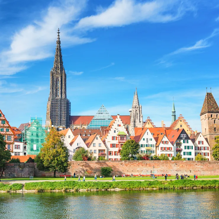 Bunte Häuser mit Giebeldächern am Fluss, dahinter Kirchtürme und Bäume.
