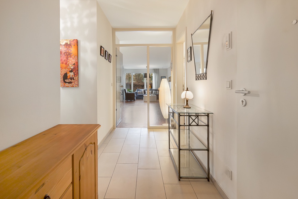 Heller Flur mit Fliesenboden, Spiegel, Glas-Konsole und Blick in ein Wohnzimmer.