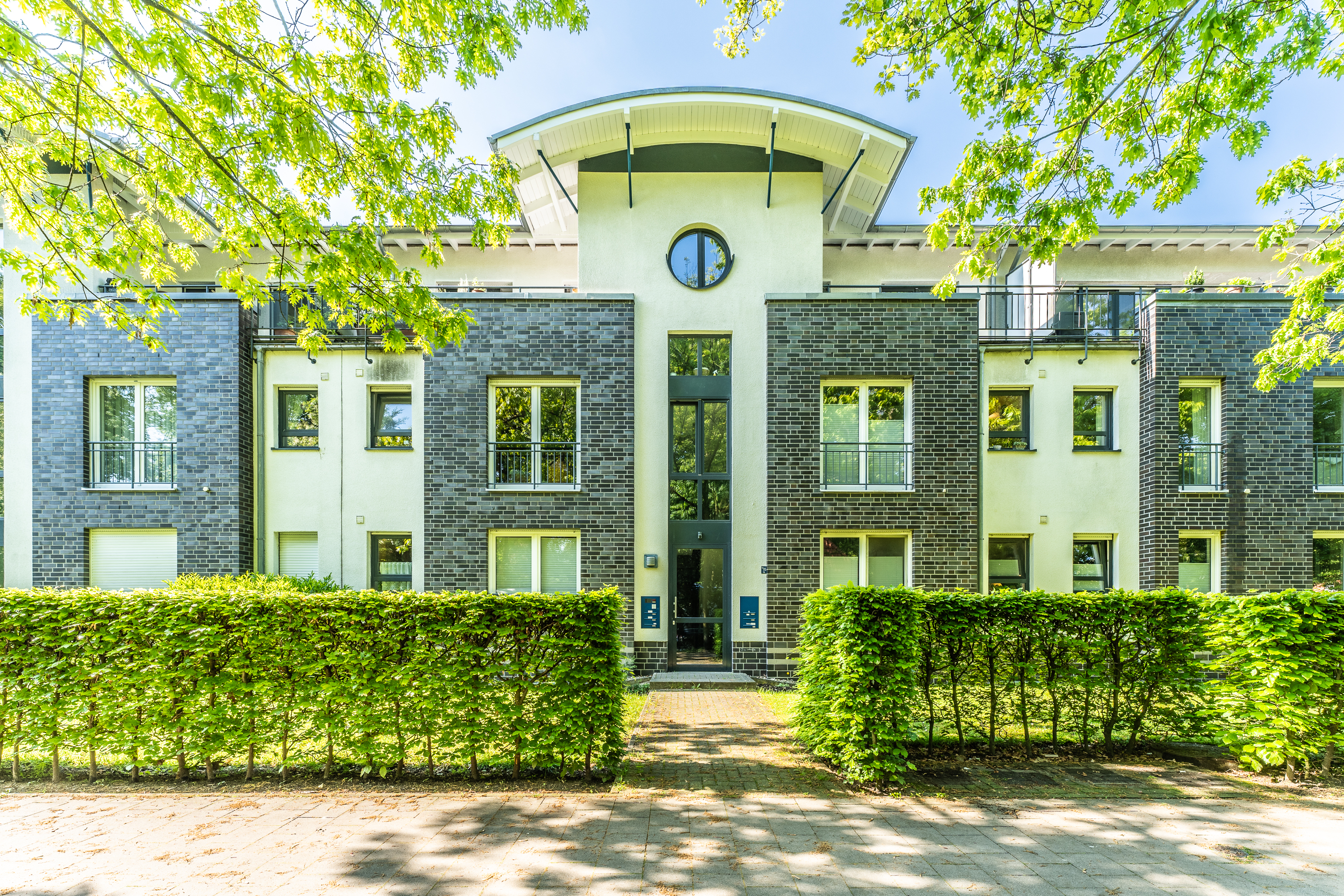 Modernes Wohngebäude mit grauer Fassade, großen Fenstern und grüner Hecke im Vordergrund.