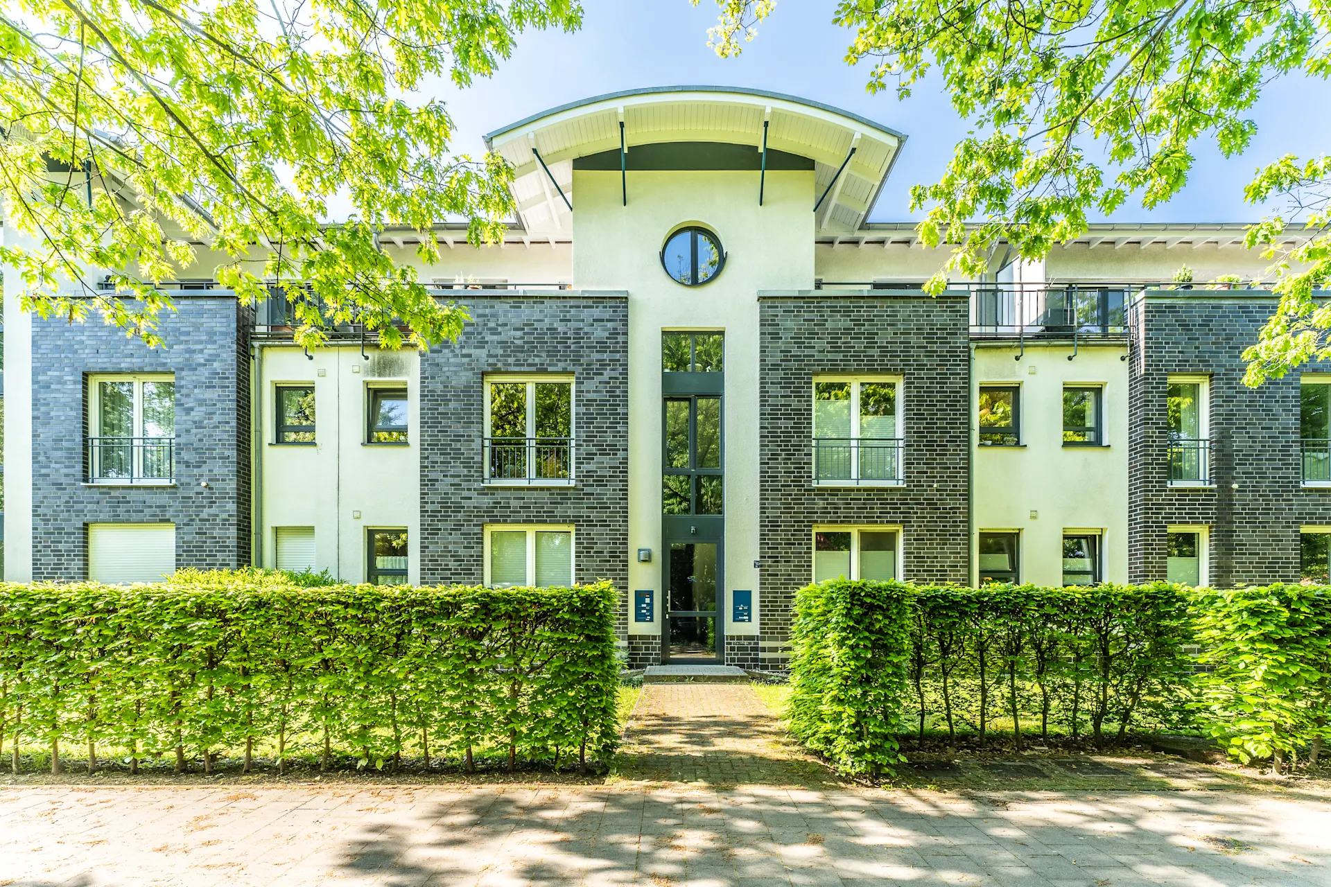 Modernes Wohngebäude mit grauer Fassade, großen Fenstern und grüner Hecke im Vordergrund.