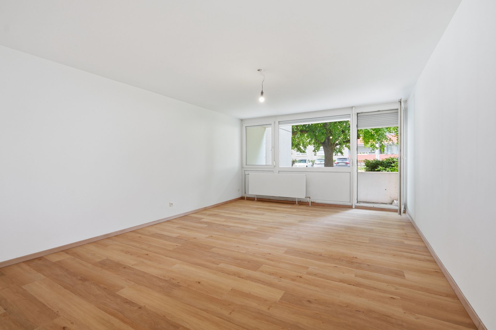 Leerer Raum mit Holzfußboden, weißen Wänden und großen Fenstern, die auf einen Baum blicken.