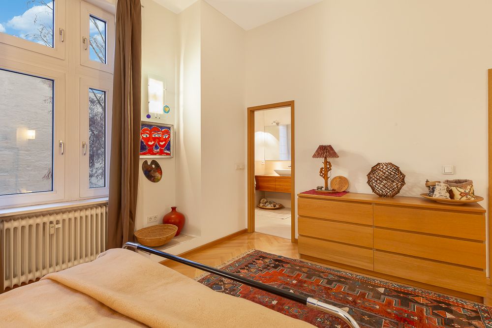 Helles Schlafzimmer mit großem Fenster, bunter Teppich, Holzkommode und Blick ins Badezimmer.