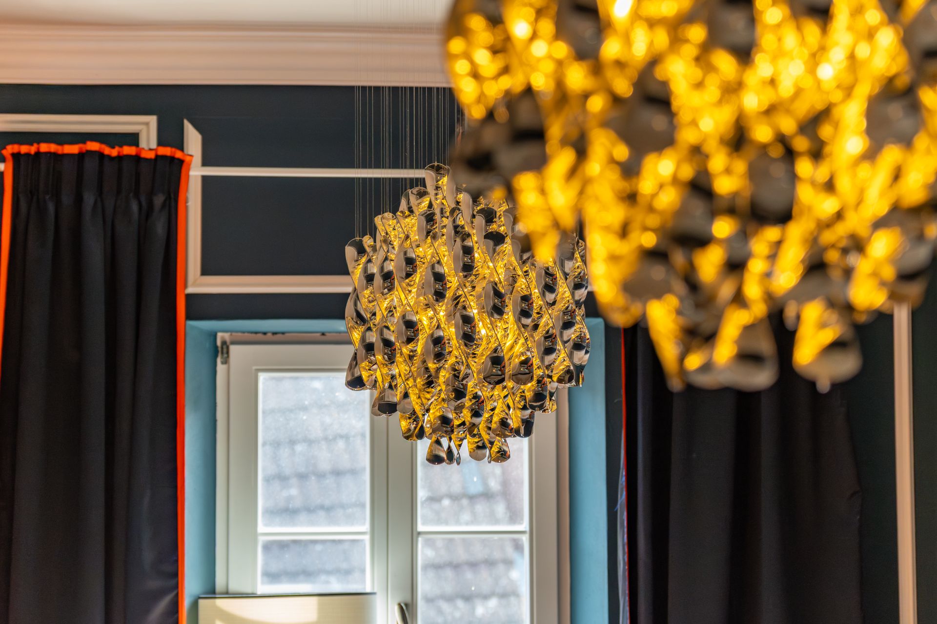 Zwei leuchtende Kronleuchter vor einem Fenster mit dunklen Vorhängen.
