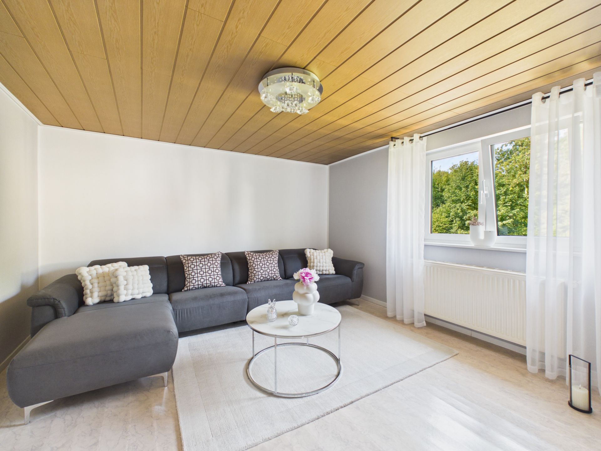Helles Wohnzimmer mit grauem Sofa, rundem Tisch und großem Fenster mit weißen Vorhängen.