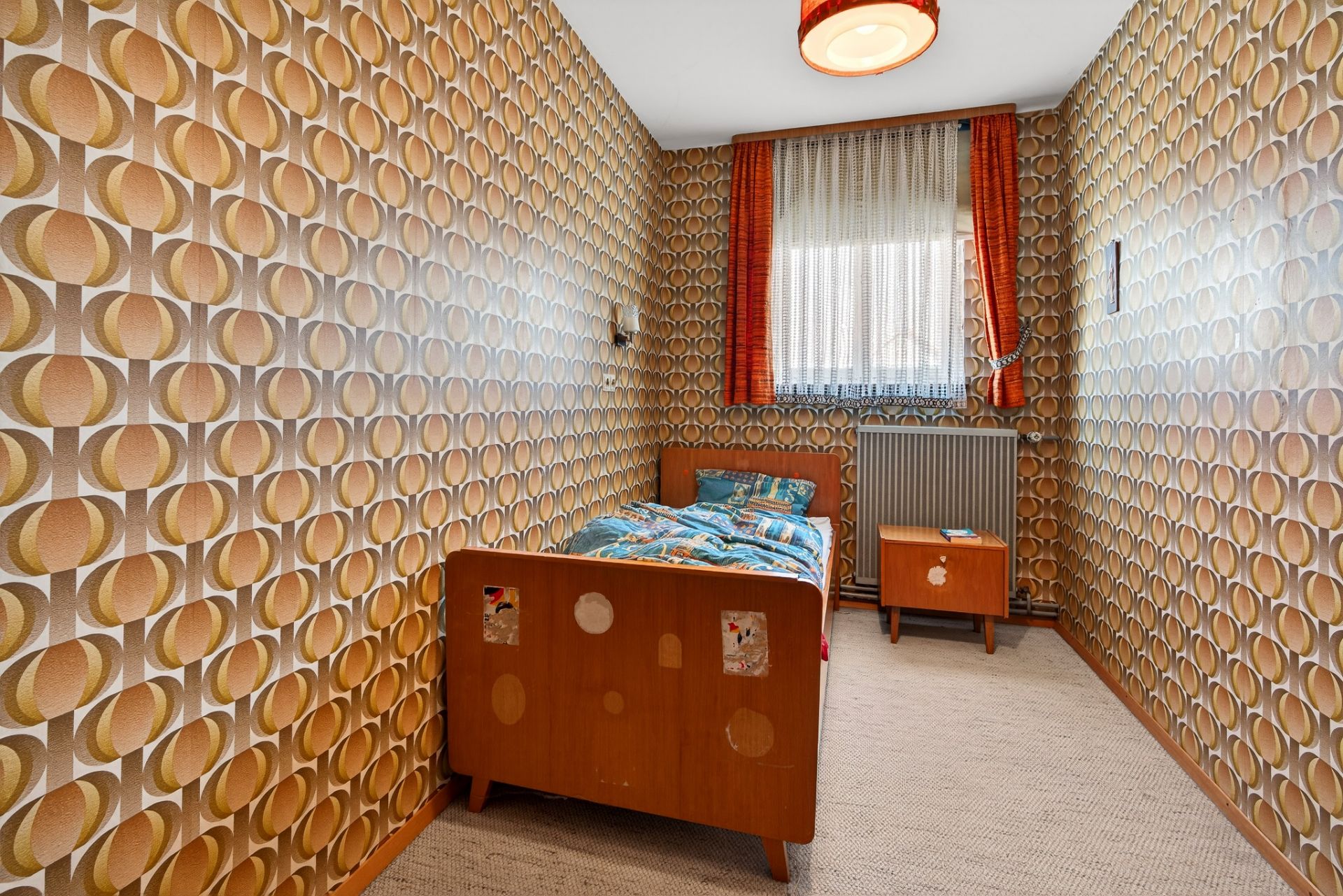 Kleines Zimmer mit Einzelbett, Nachttisch und auffälliger Retro-Tapete mit Kreis-Muster.
