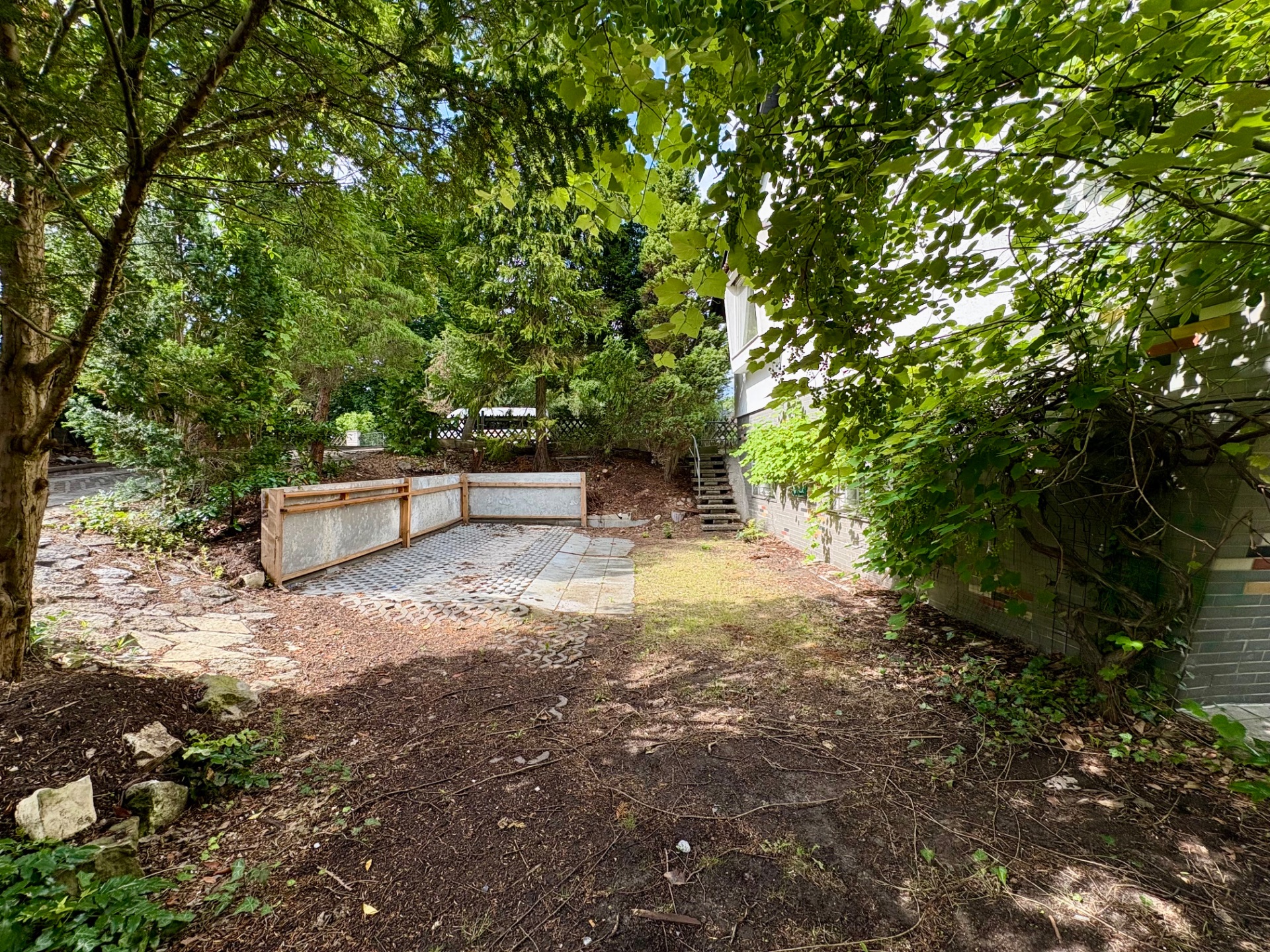 Gartenbereich mit Bäumen, befestigtem Platz und kleiner Treppe am Haus.