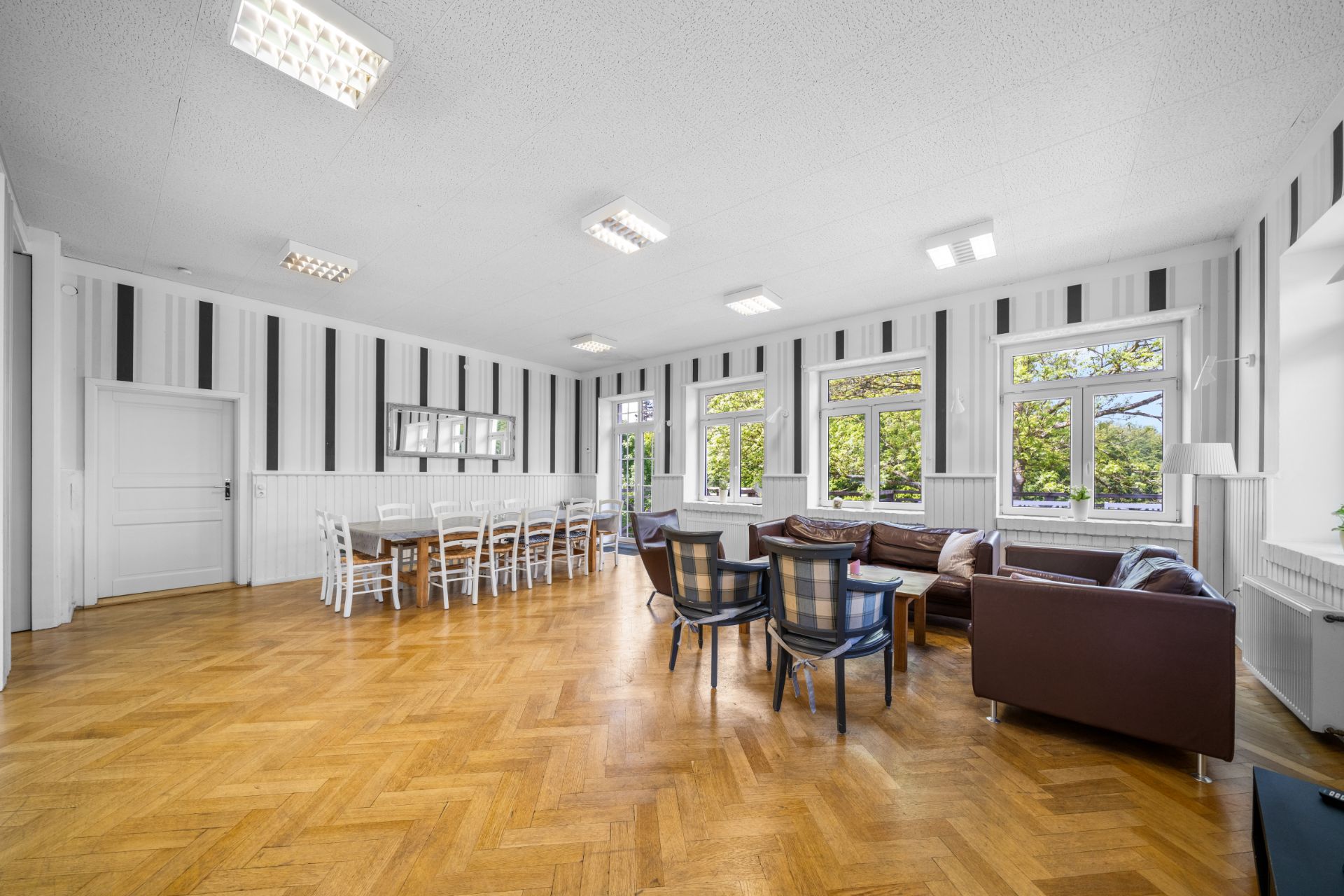 Heller Wohnraum mit Esstisch, Stühlen, Sofas und großen Fenstern, Parkettboden.