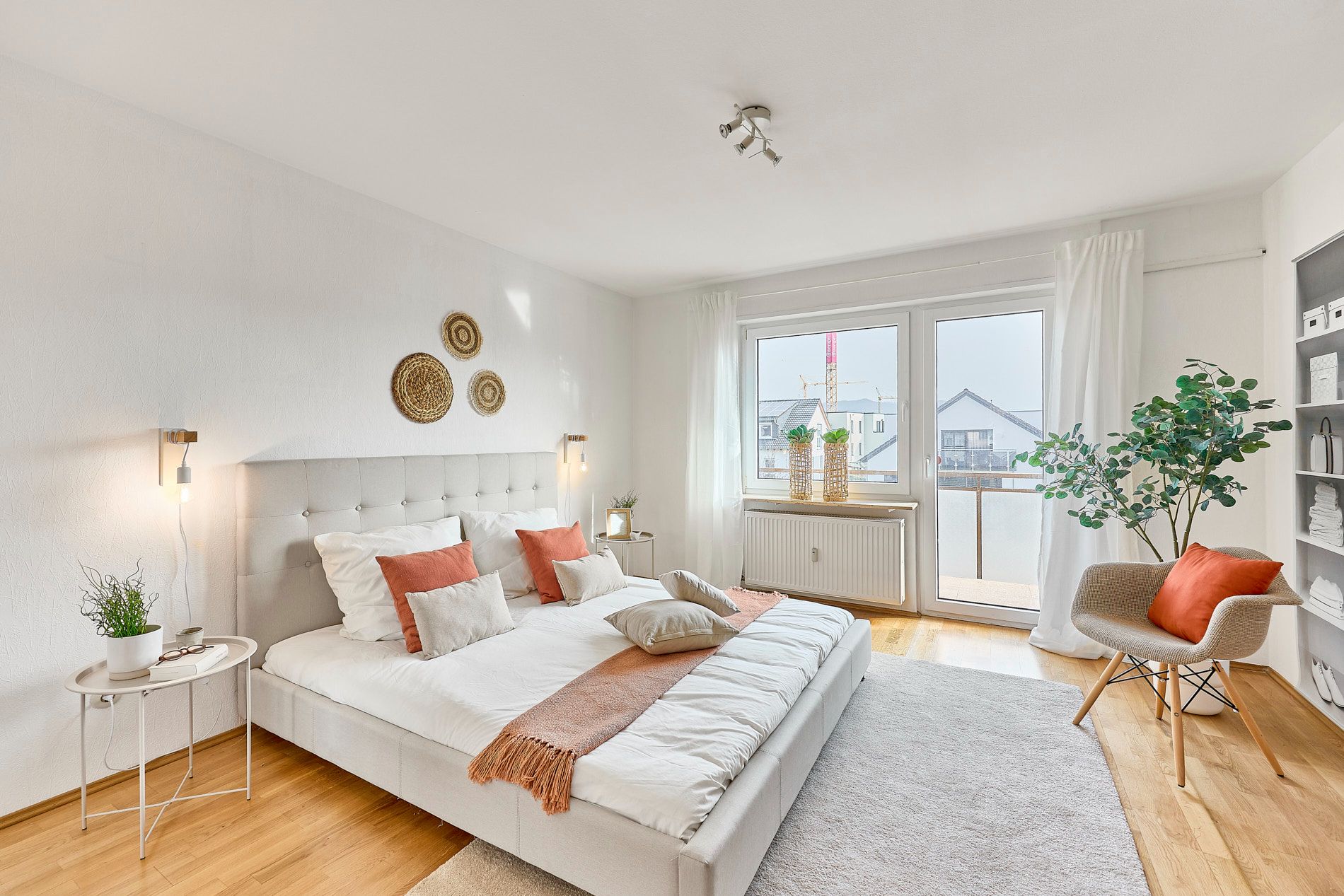 Helles Schlafzimmer mit Doppelbett, Dekokissen, Pflanzen, Sessel und großem Fenster mit Balkon.