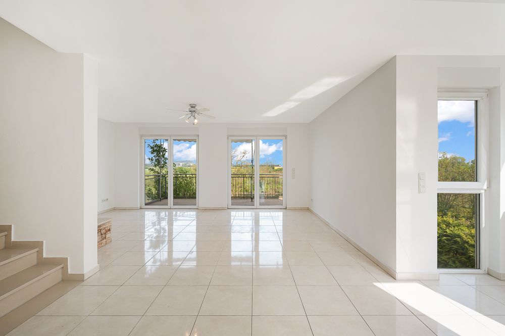Helles Wohnzimmer mit Fliesenboden, großen Fenstern und Zugang zum Balkon, Treppe links im Bild.