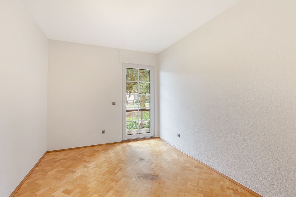 Leeres Zimmer mit Parkettboden und Glastür, die nach draußen führt.