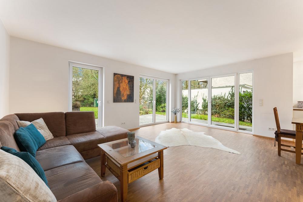 Helles Wohnzimmer mit großer Eckcouch, Glastisch und bodentiefen Fenstern zum Garten.