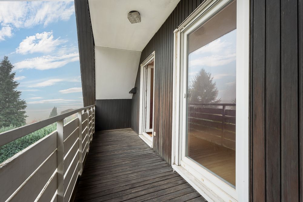 Langer Balkon mit Holzboden, weißen Geländern und zwei großen Glastüren, Blick ins Grüne.