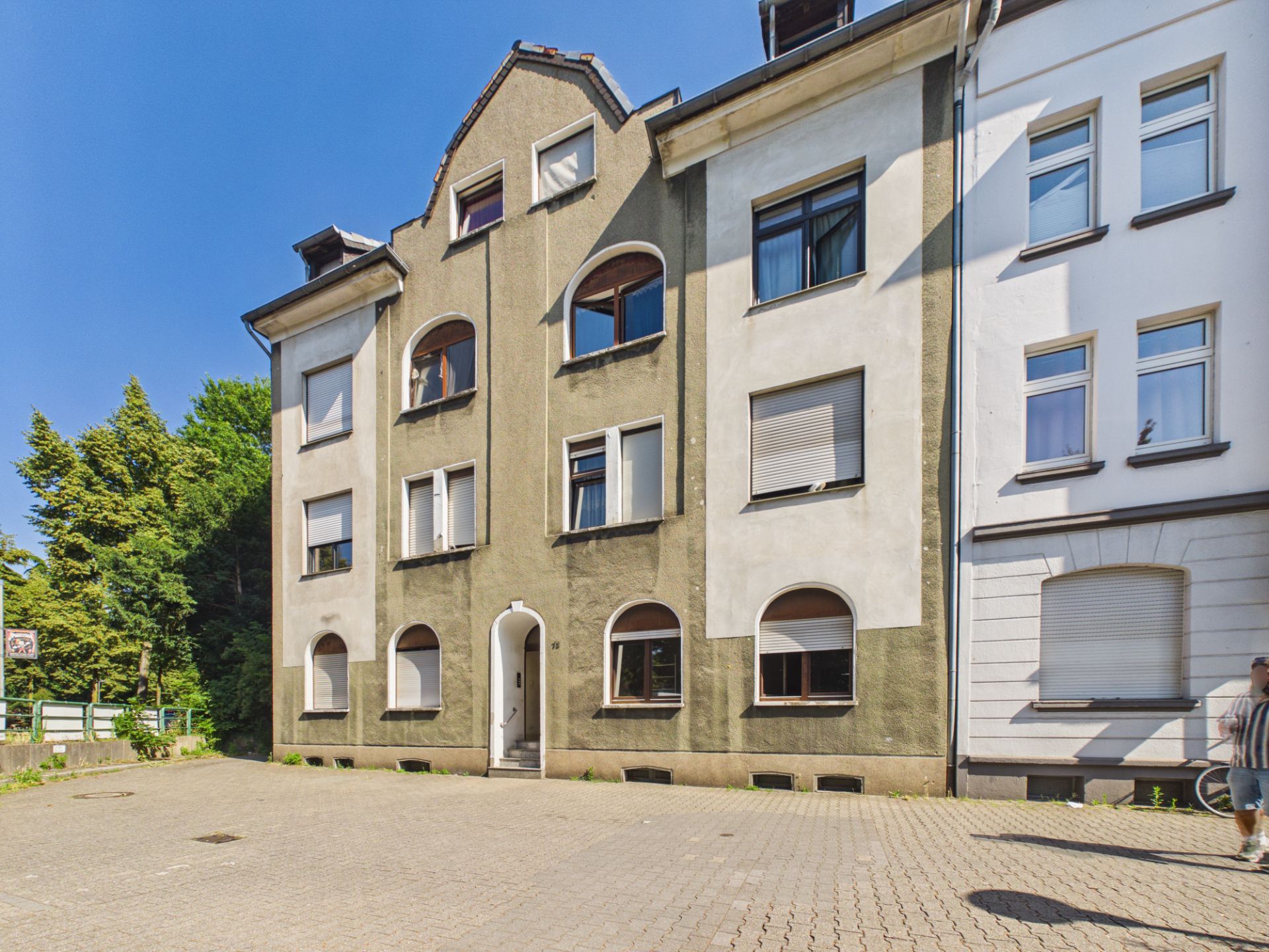 Mehrfamilienhaus mit grauer Fassade, mehrere Fenster und Eingangsbereich, gepflasterter Vorplatz.