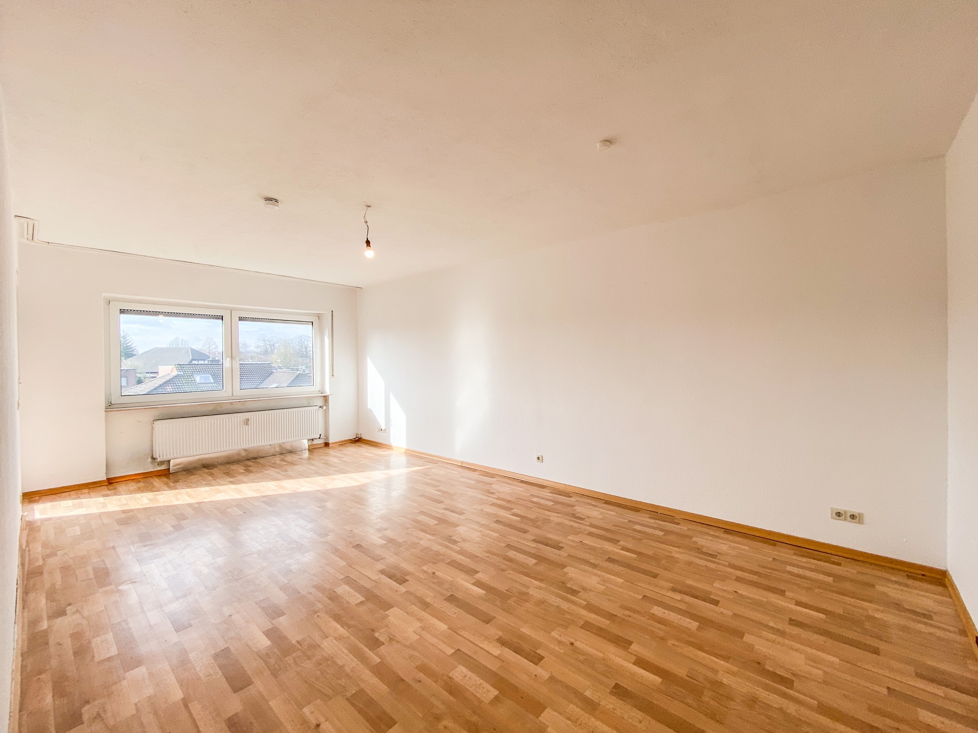 Heller, leerer Raum mit Parkettboden, weißen Wänden und zwei großen Fenstern.