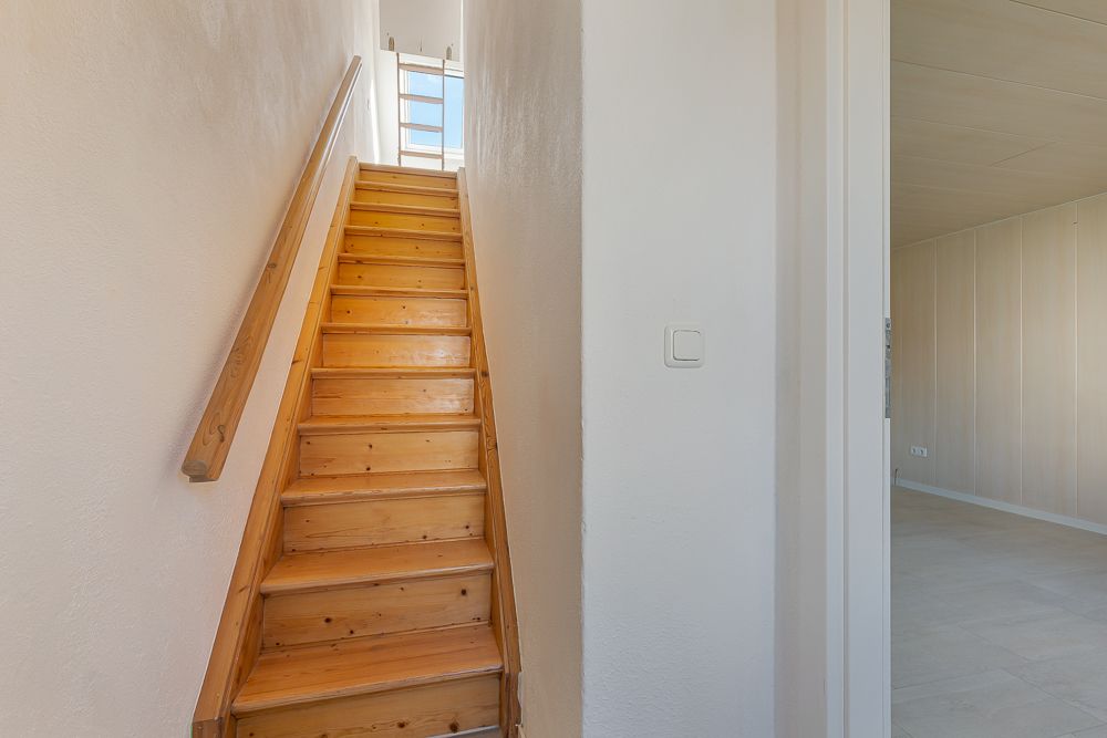 Holztreppe mit Handlauf führt nach oben, daneben ein heller Raum mit weißen Wänden.