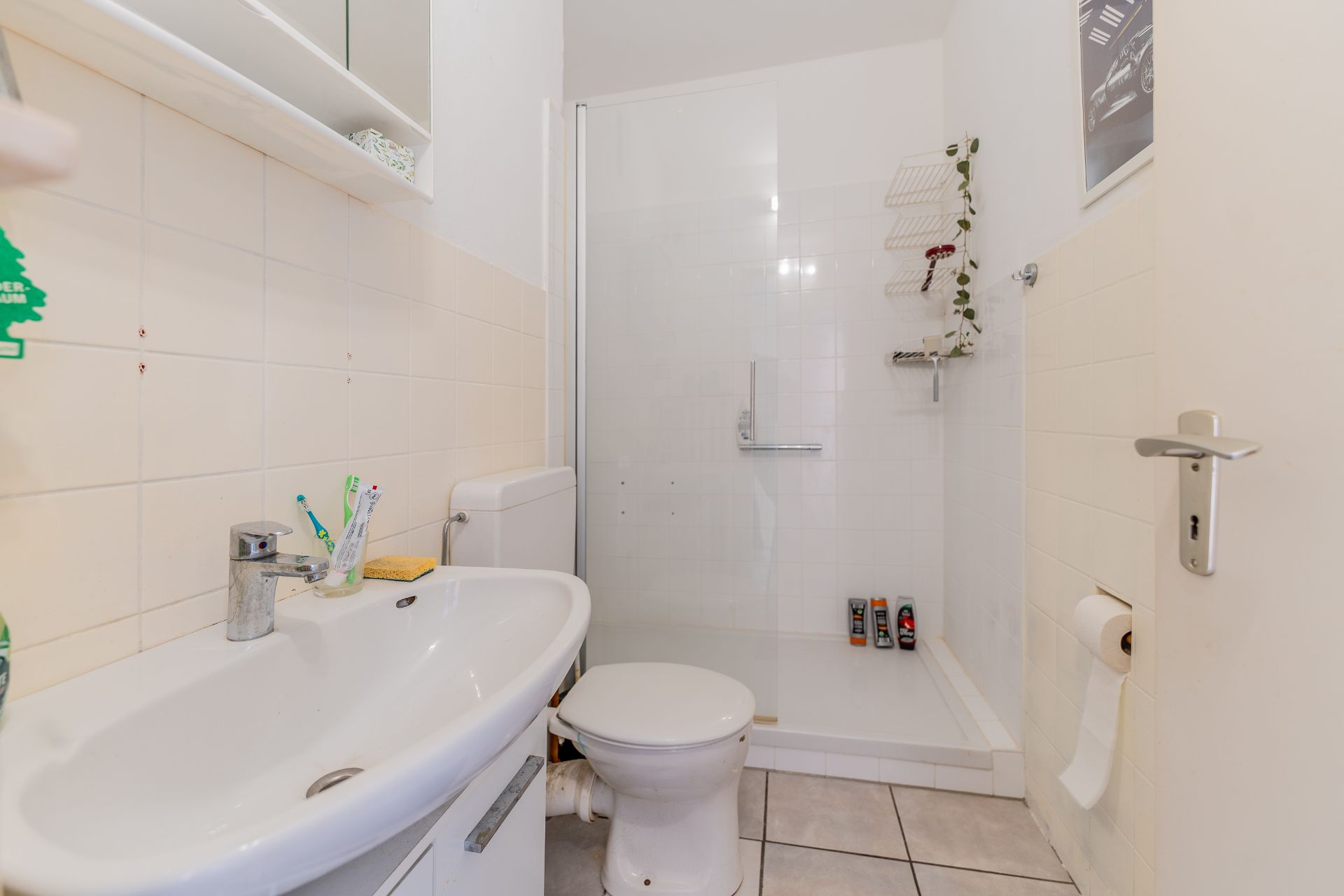 Badezimmer mit Waschbecken, Toilette und ebenerdiger Dusche, weiße Fliesen und Ablageflächen.