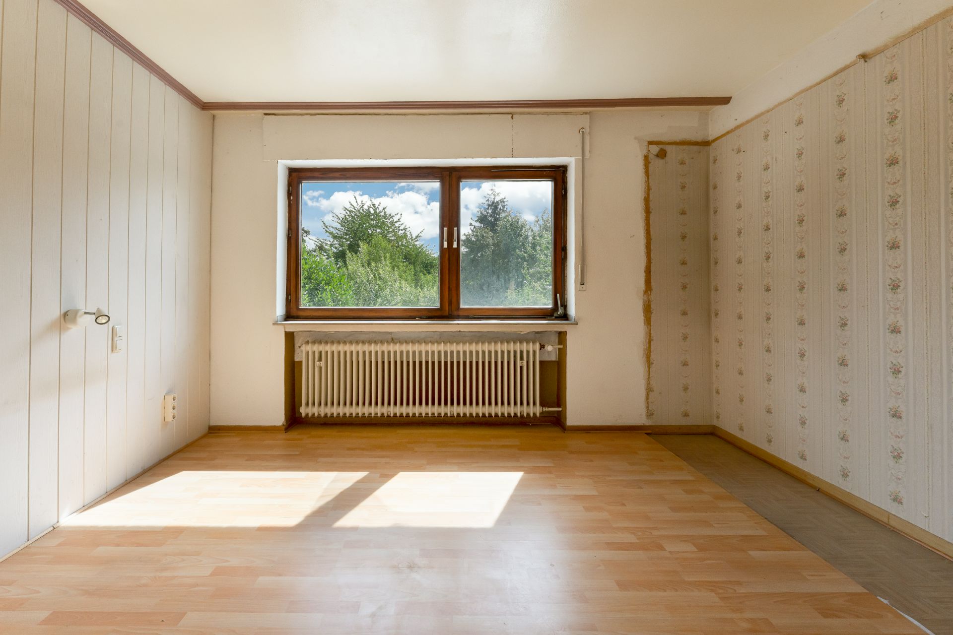 Leerer Raum mit großem Fenster, Heizkörper und hellem Laminatboden.