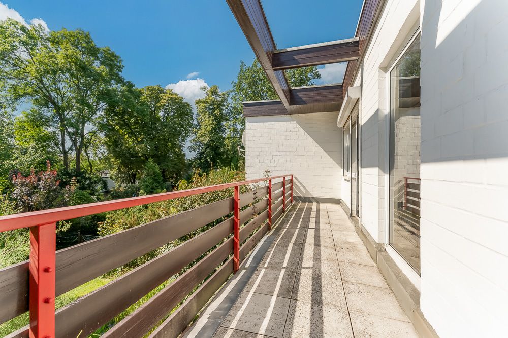 Heller Balkon mit rotem Geländer und seitlichem Zugang, daneben grüne Bäume im Garten.