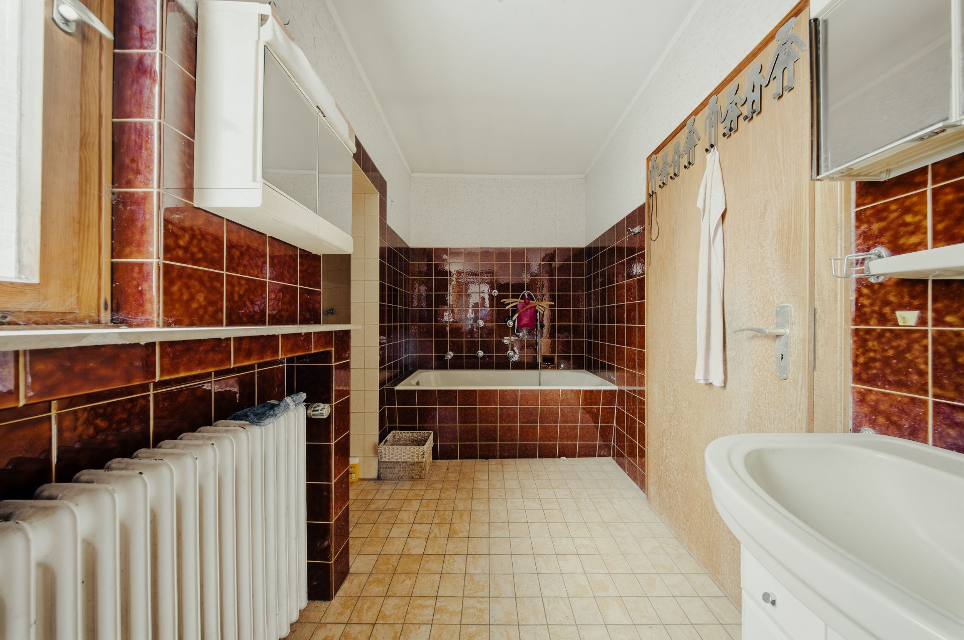 Badezimmer mit braunen Fliesen, Badewanne, Waschbecken, Heizkörper und Fenster.