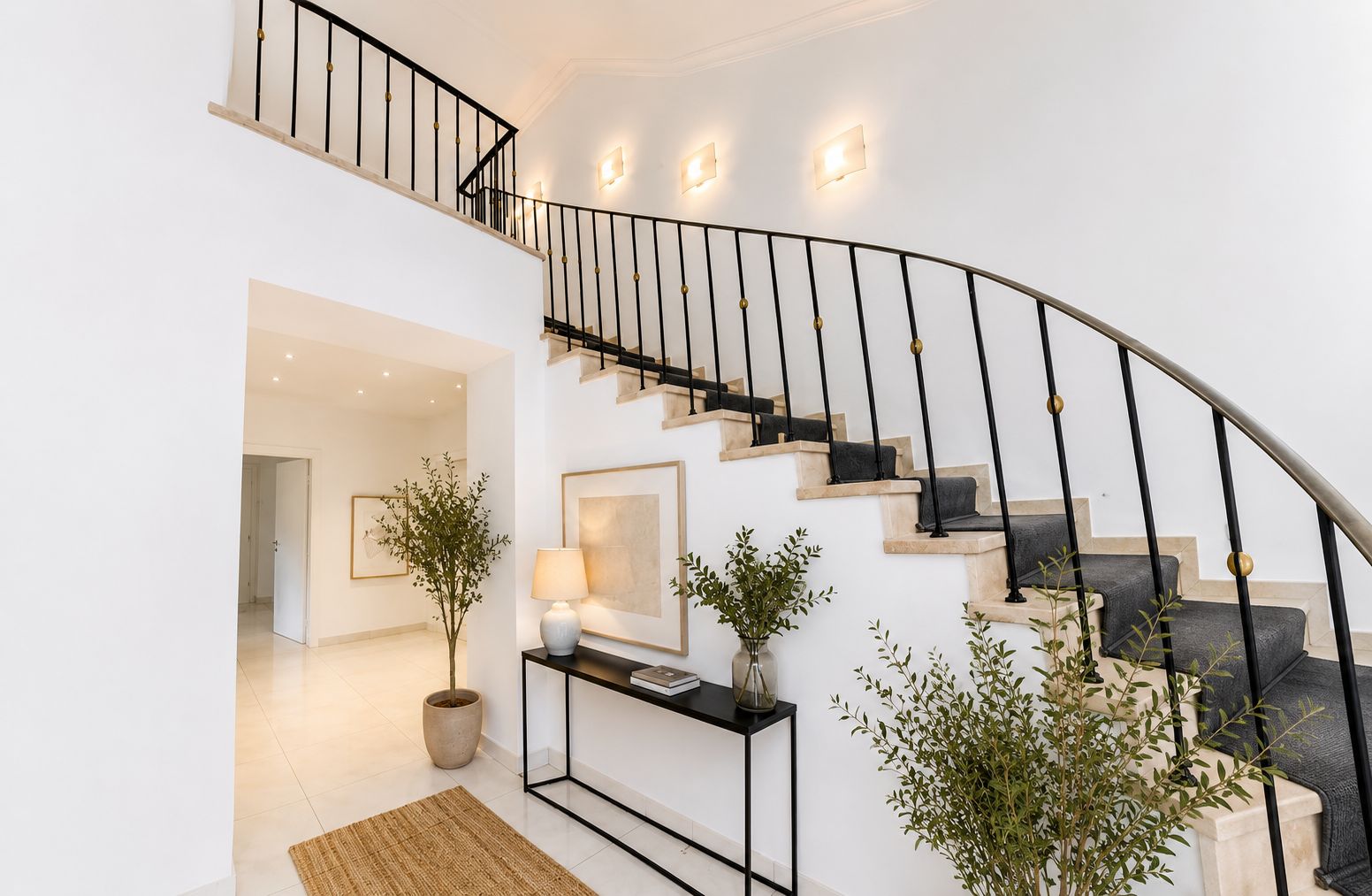 Heller Eingangsbereich mit breiter Treppe, Pflanzen, Konsole und Wandlampen.