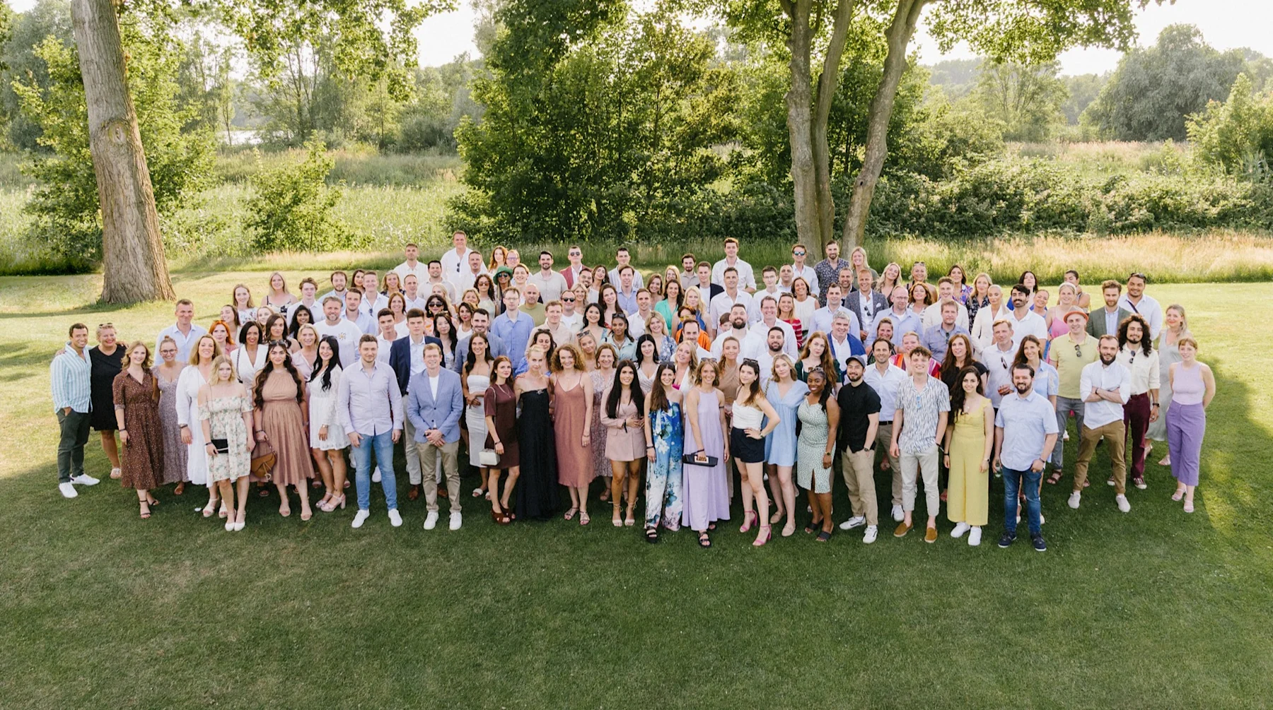 Eine große Gruppe von Menschen steht im Freien auf einer Wiese, Bäume im Hintergrund.