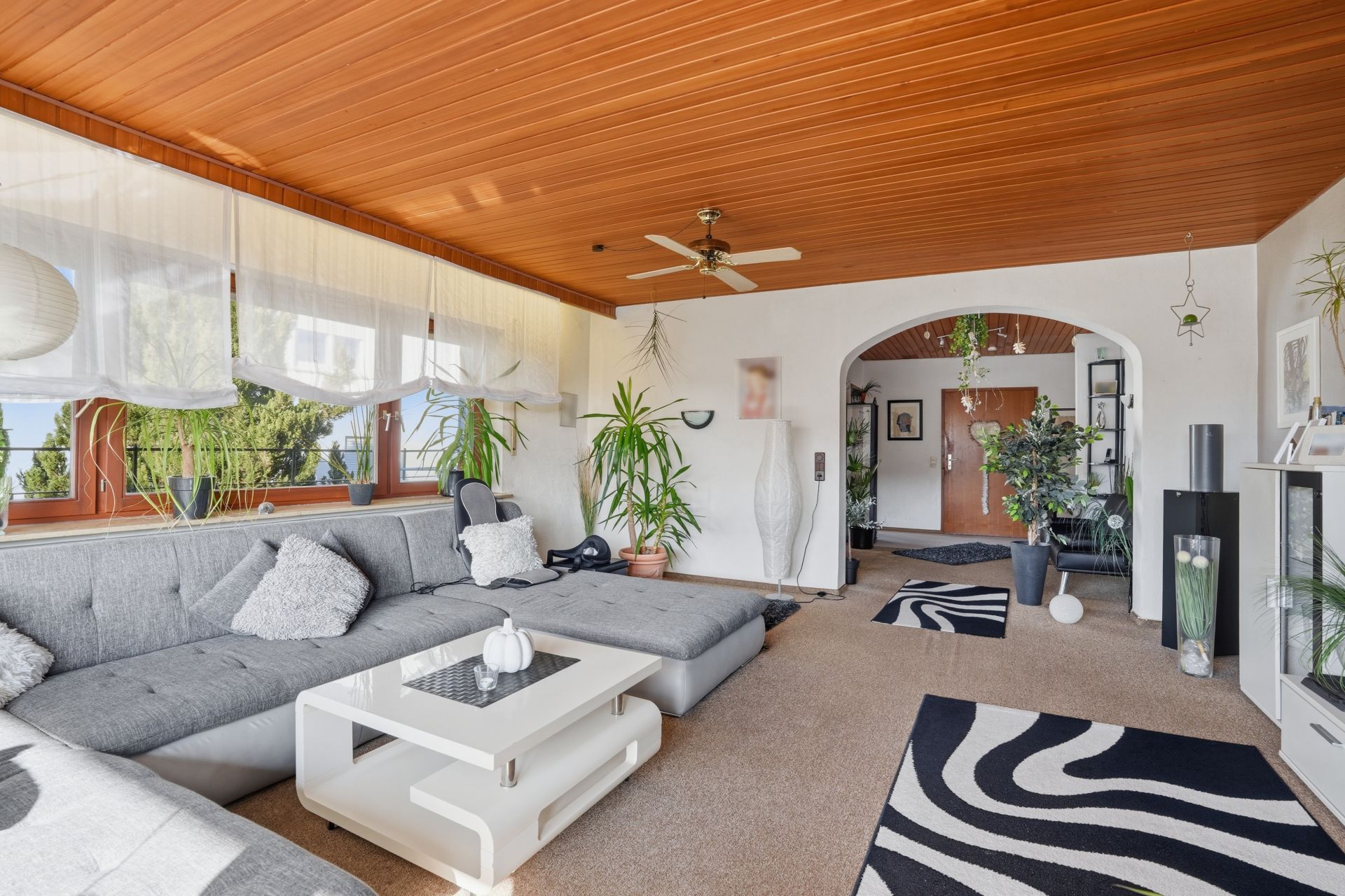 Helles Wohnzimmer mit grauem Sofa, Holzbalkendecke und vielen Zimmerpflanzen.