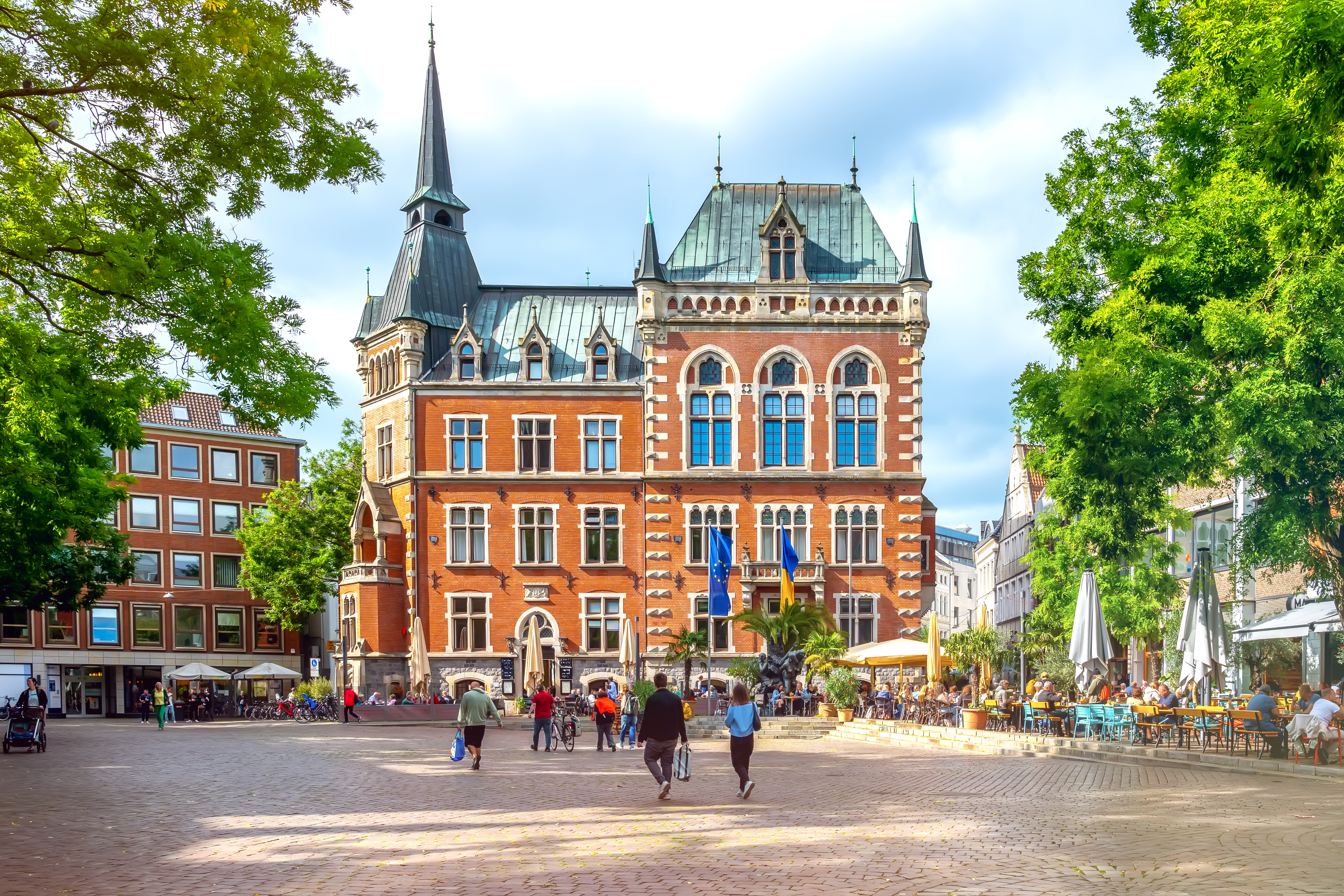 Historisches Backsteingebäude mit Türmen auf einem belebten Platz, umgeben von Bäumen und Cafés.