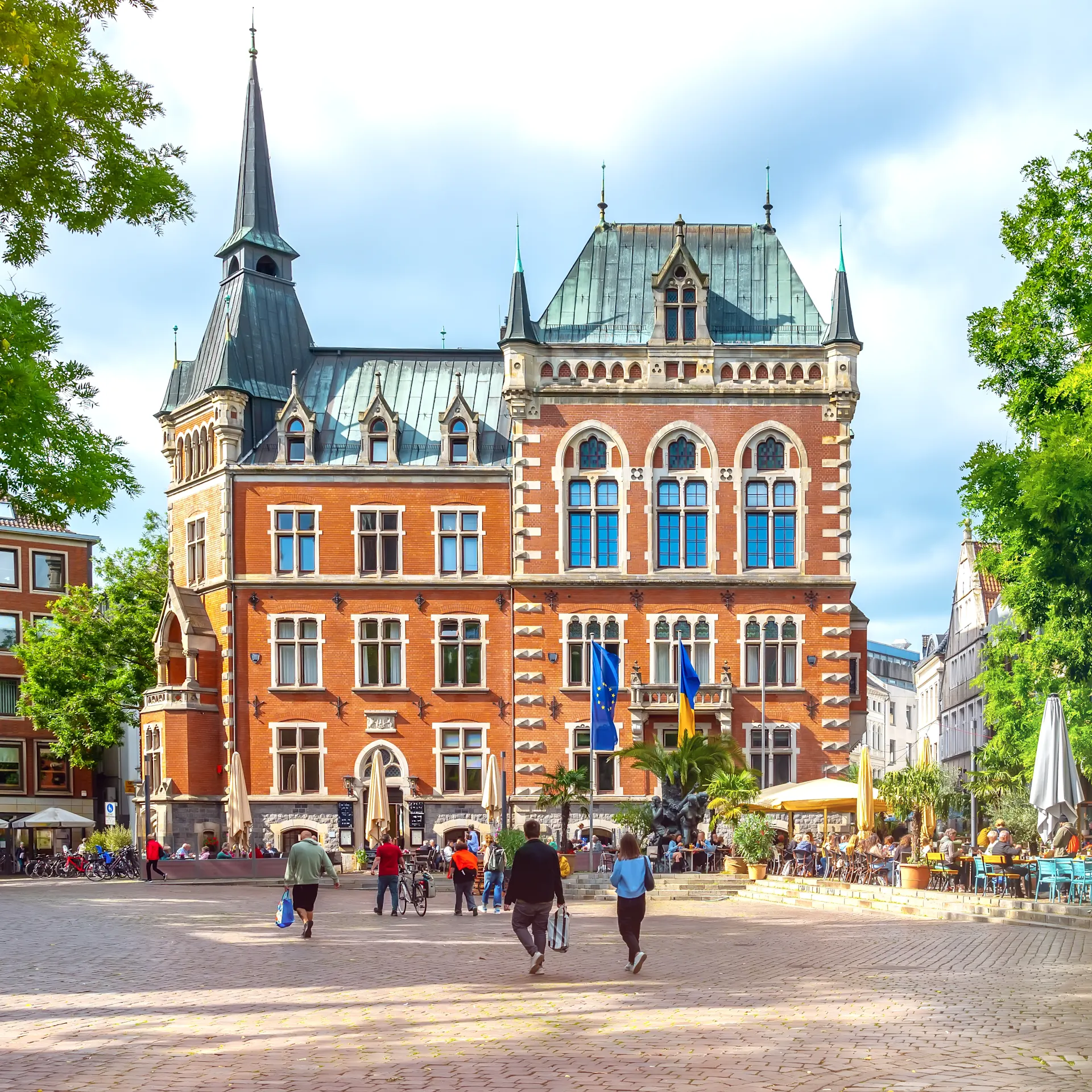 Historisches Backsteingebäude mit Türmen auf einem belebten Platz, umgeben von Bäumen und Cafés.