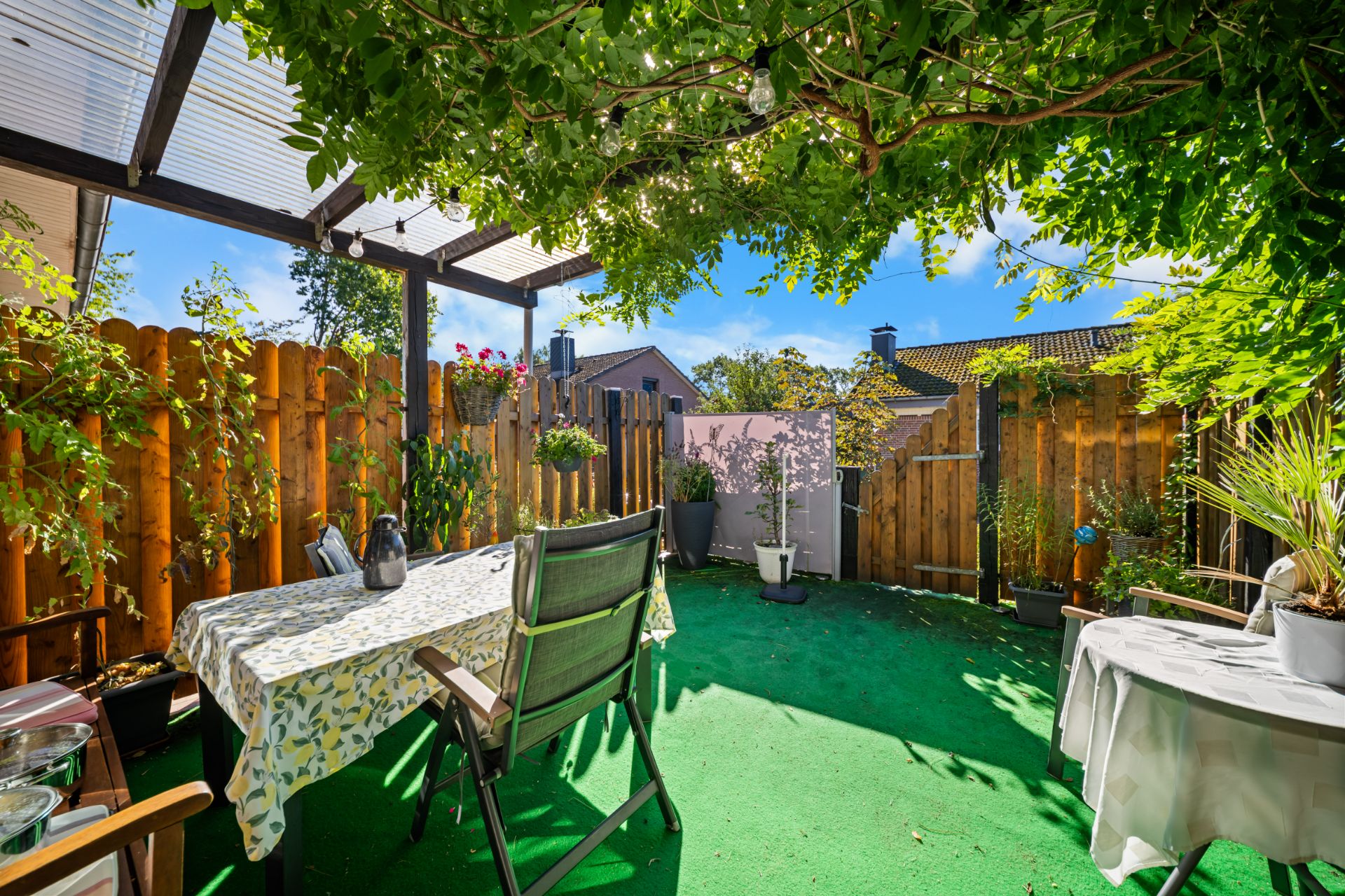 Überdachte Terrasse mit grünem Boden, Tisch, Stühlen und vielen Pflanzen vor Holzzaun.