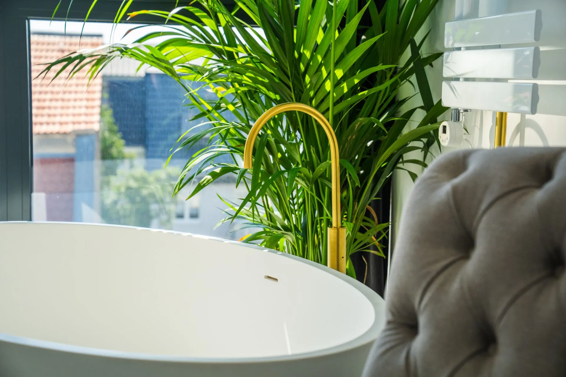 Freistehende Badewanne mit goldener Armatur, großer Pflanze und Fenster im Hintergrund.