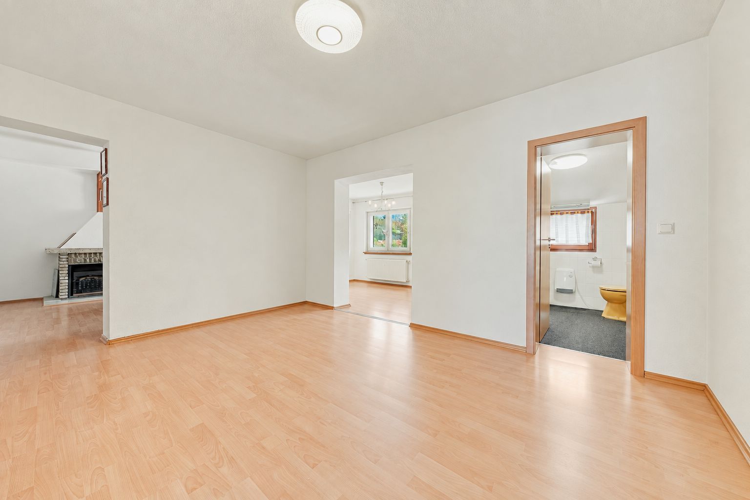 Helles Wohnzimmer mit Parkettboden, weißen Wänden, Tür zum Bad und Zugang zu weiteren Räumen.