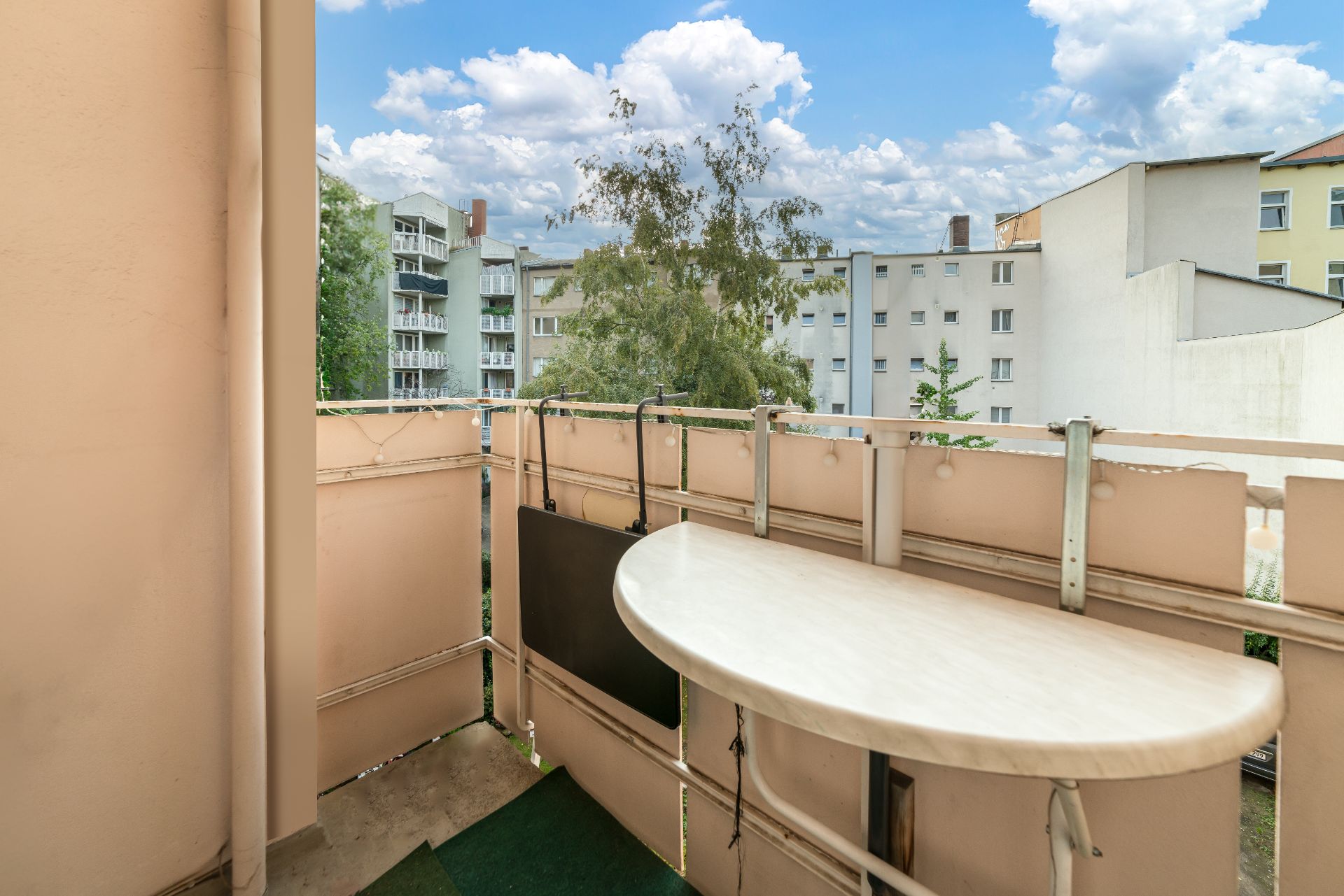 Kleiner Balkon mit Tisch, Blick auf Nachbarhäuser und Bäume bei Tageslicht.