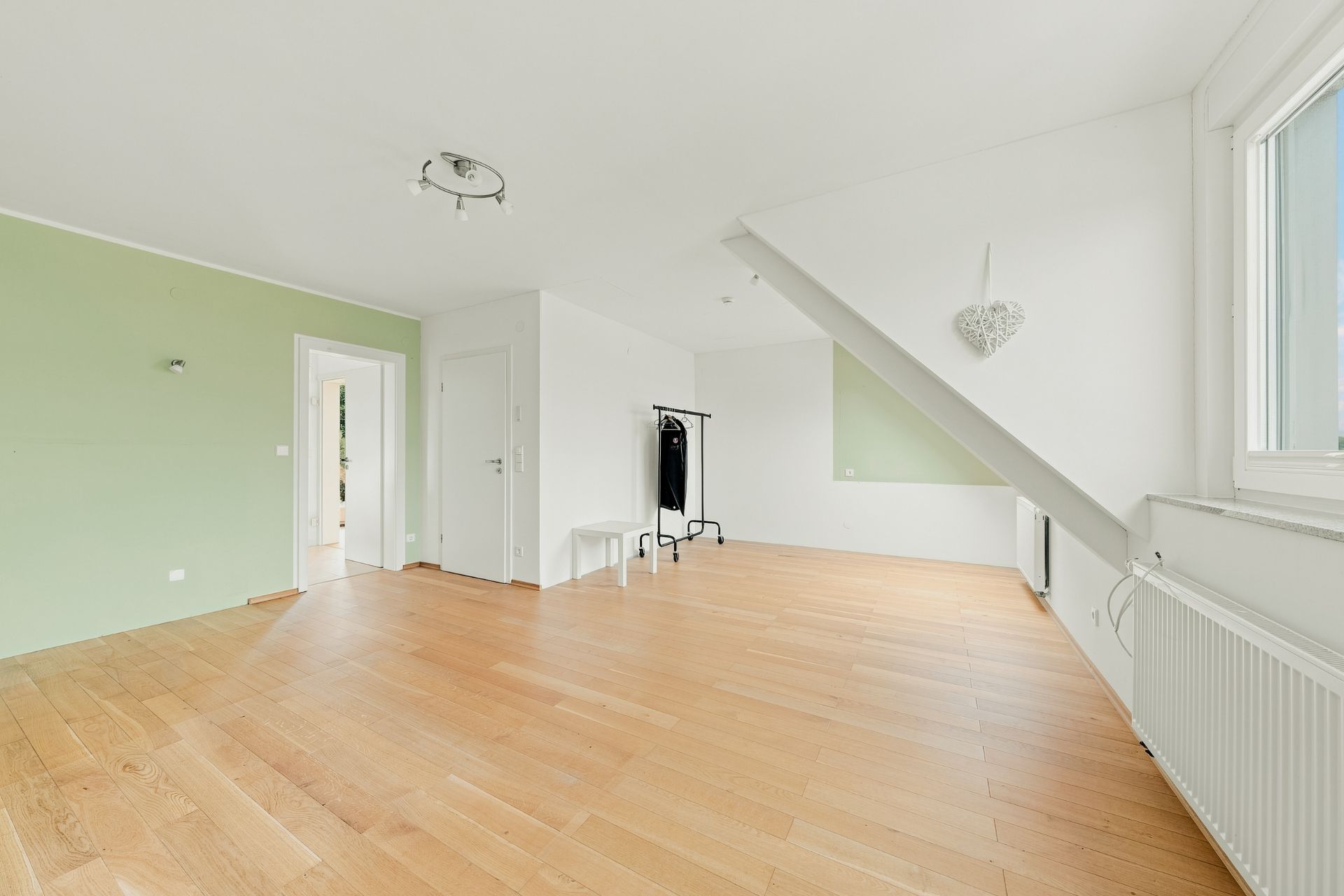 Heller Raum mit Holzfußboden, grün-weißen Wänden, Fenster, Heizkörper und Kleiderständer.