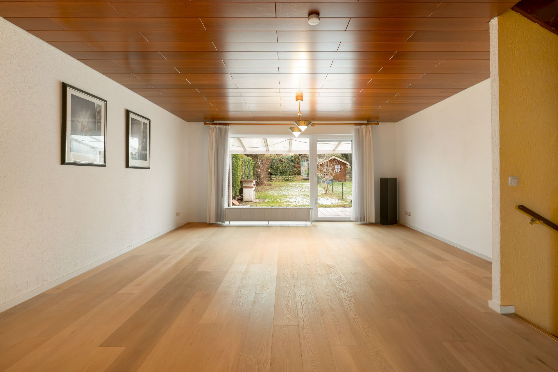 Helles Wohnzimmer mit Holzfußboden, Deckenlampe und großen Fenstern zum Garten.