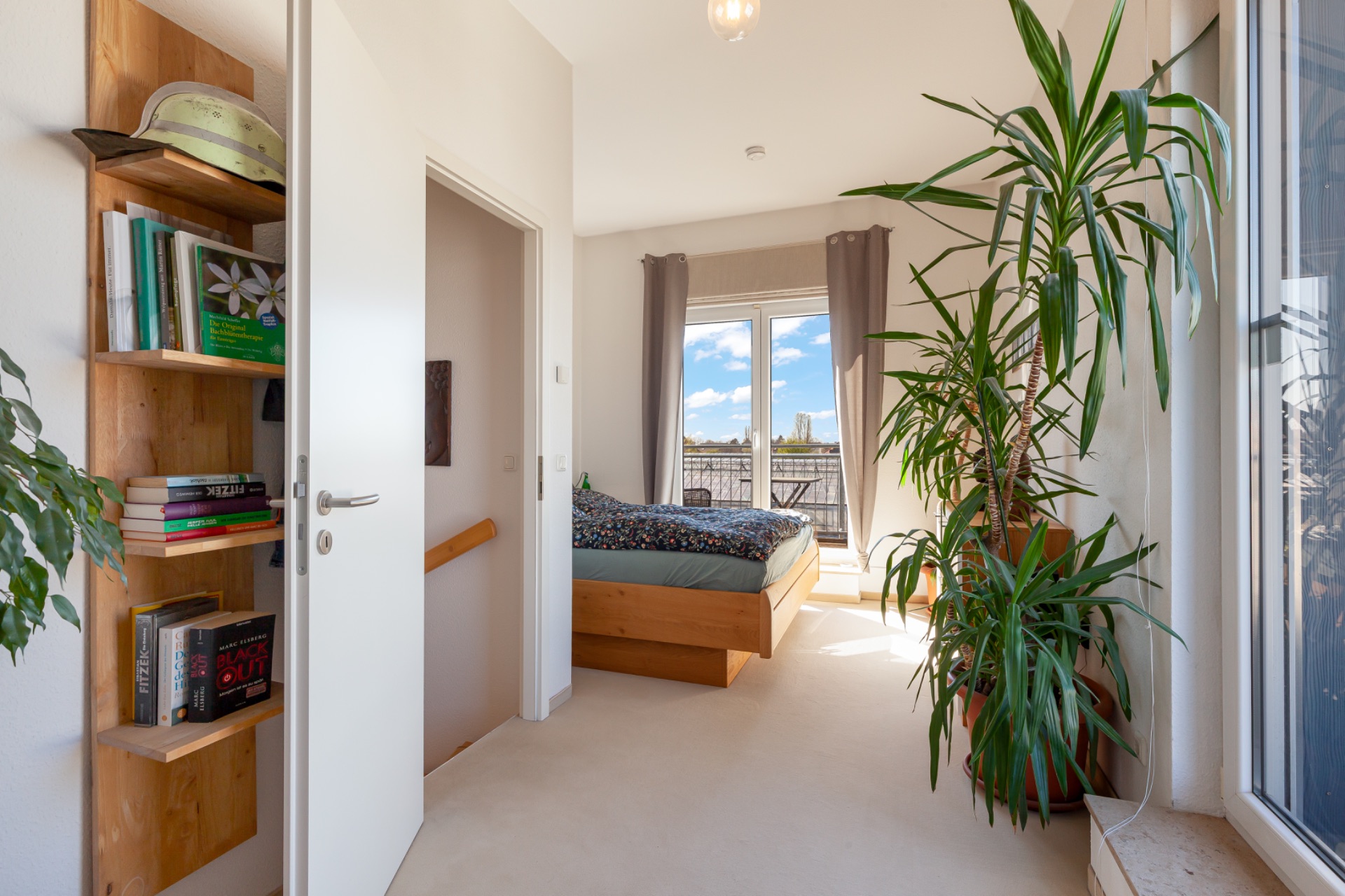 Helles Schlafzimmer mit großem Fenster, Bett, Regal und mehreren grünen Pflanzen.