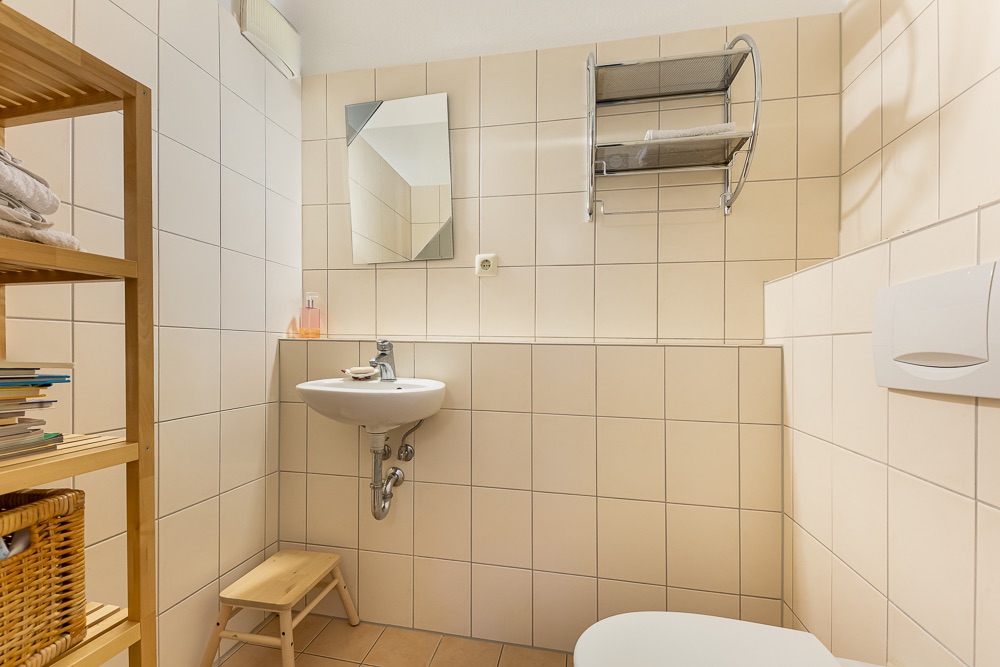 Kleines Bad mit Waschbecken, Spiegel, Regal, Hocker und Wandregal mit Handtüchern.