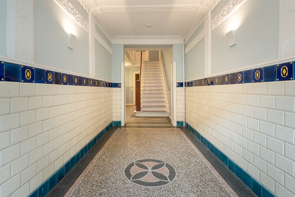 Heller Flur mit blauen Fliesen, verzierten Wänden und Treppe am Ende.
