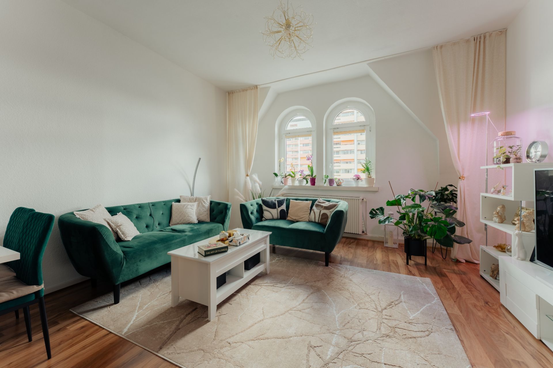 Helles Wohnzimmer mit zwei grünen Sofas, Couchtisch, Teppich und Zimmerpflanzen vor zwei Fenstern.