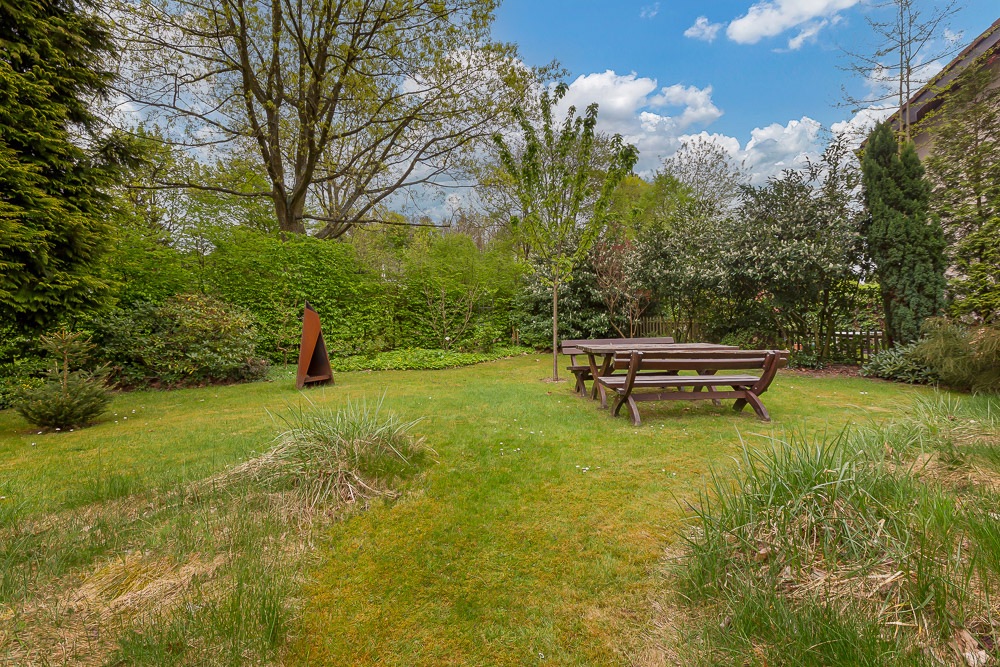 Grüner Garten mit Holzbänken und -tisch, Bäumen und einem kleinen Kunstobjekt.