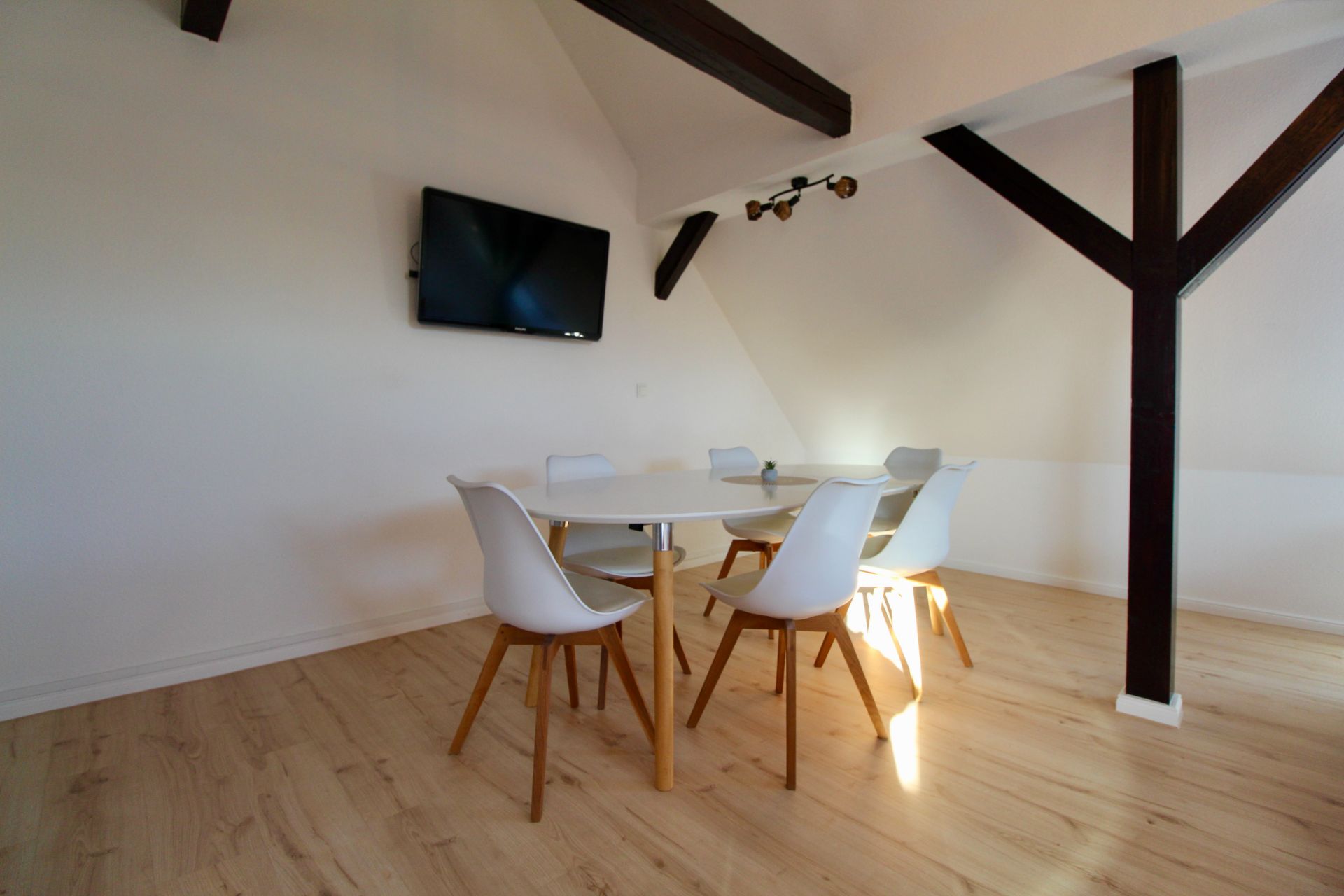 Essbereich mit rundem Tisch, sechs weißen Stühlen und Wandfernseher in einem Raum mit Holzbalken.