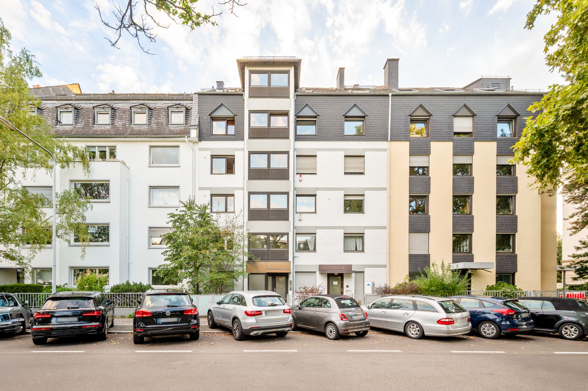 Mehrstöckiges Wohnhaus mit geparkten Autos davor und Bäumen an den Seiten.